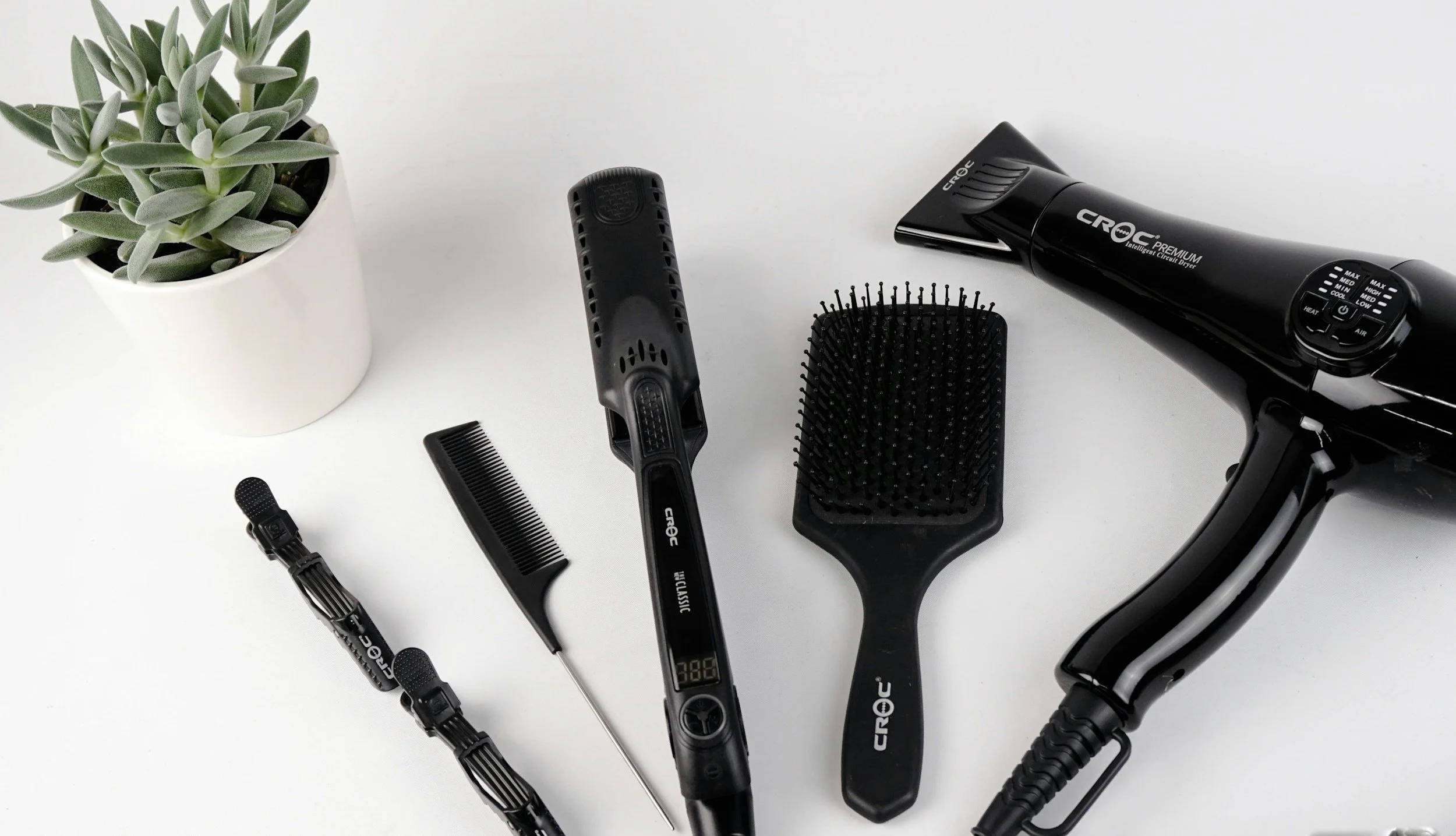 Collection of hair styling tools including a hairdryer, hairbrush, comb, flat iron, and a small black hair clip, arranged on a white surface with a potted green succulent plant.