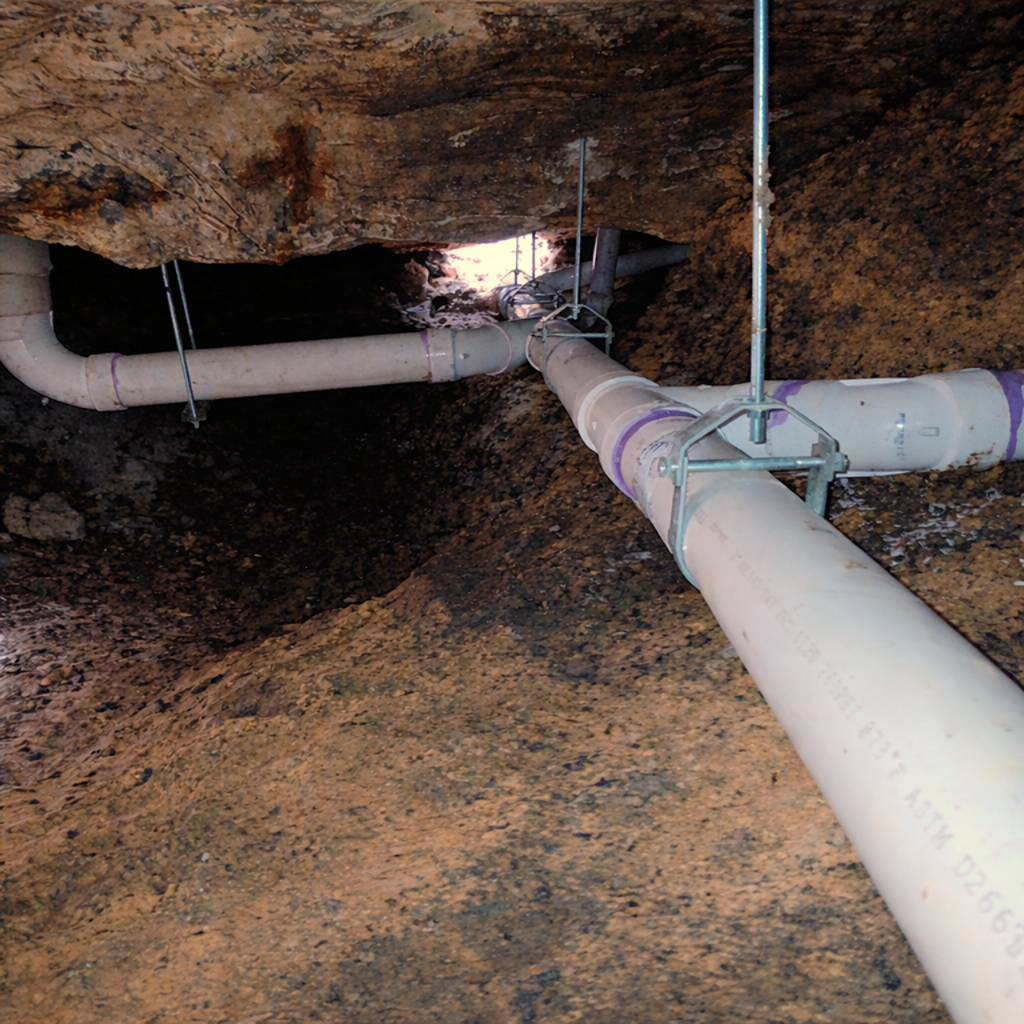 Underground pipe installation with white PVC pipes supported by metal hangers, in a rocky trench with dirt walls.