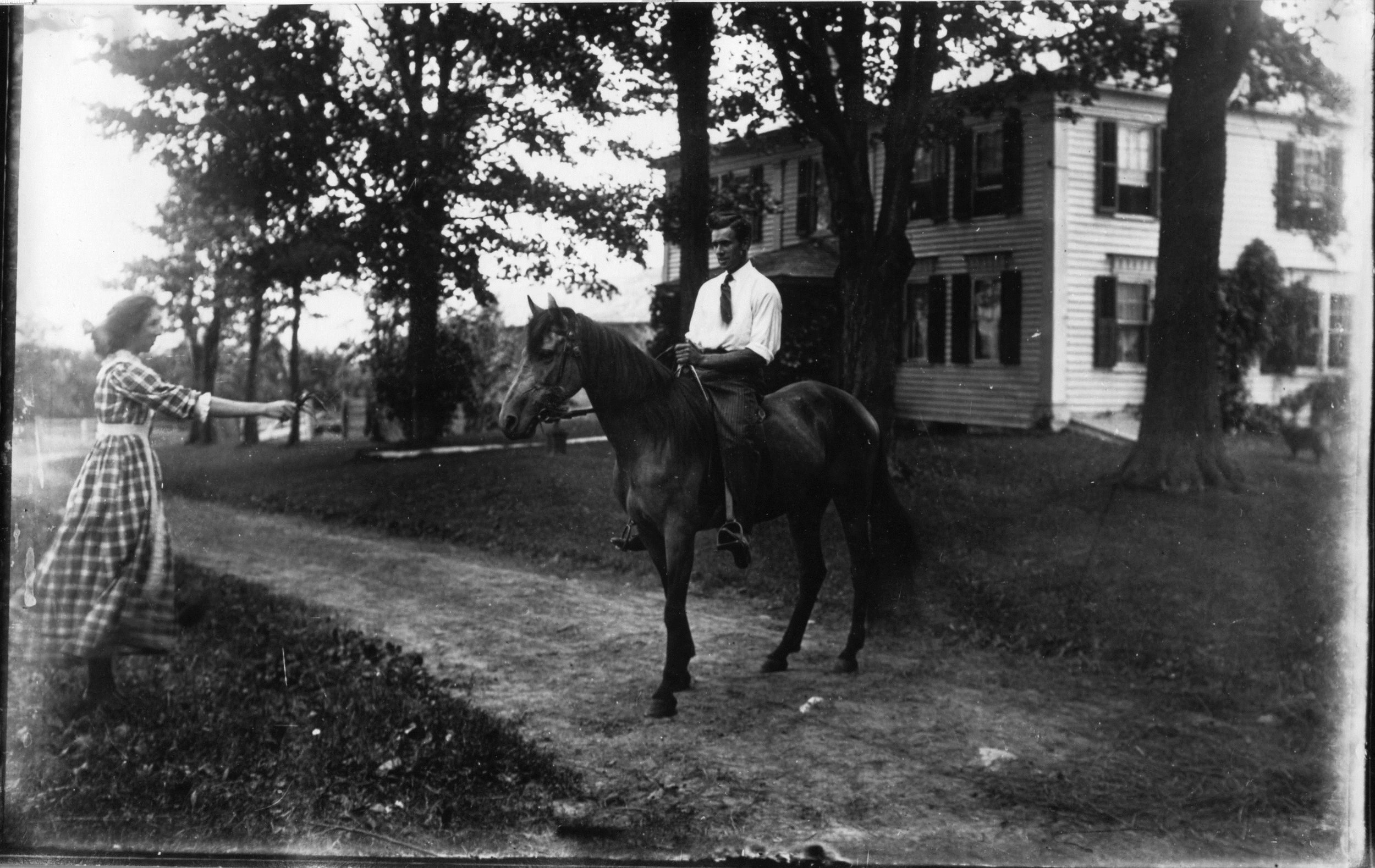 A black and white photograph shows a young man sitting on a horse, while a woman in a plaid dress stands nearby holding the horse's reins. There are large trees and a two-story house in the background.
