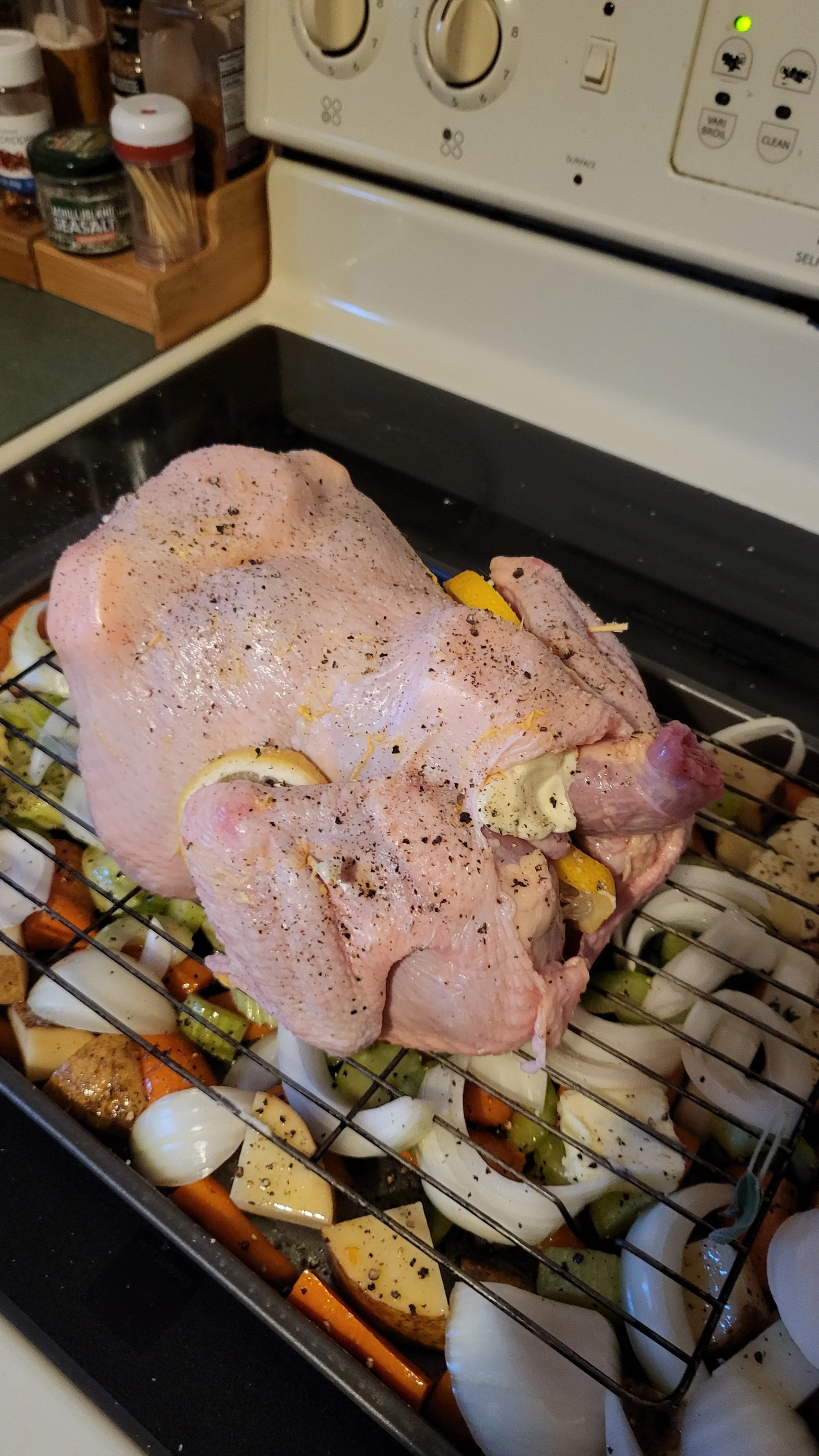 Raw whole chicken seasoned with black pepper and herbs resting on a roasting rack over cut vegetables in a roasting pan, with a stove and spice jars in the background.