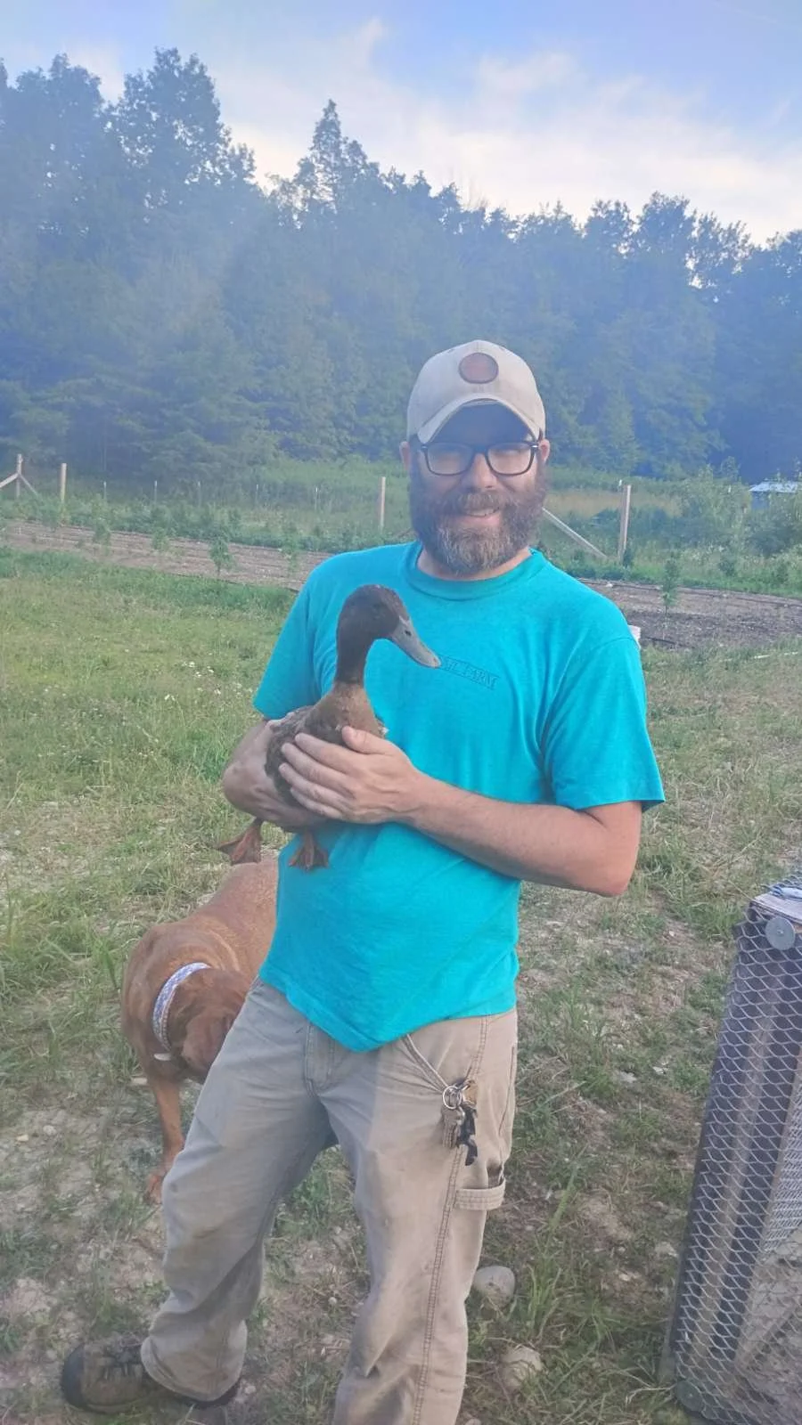 Man with glasses and a beard holding a duck outside in a grassy area with trees in the background.