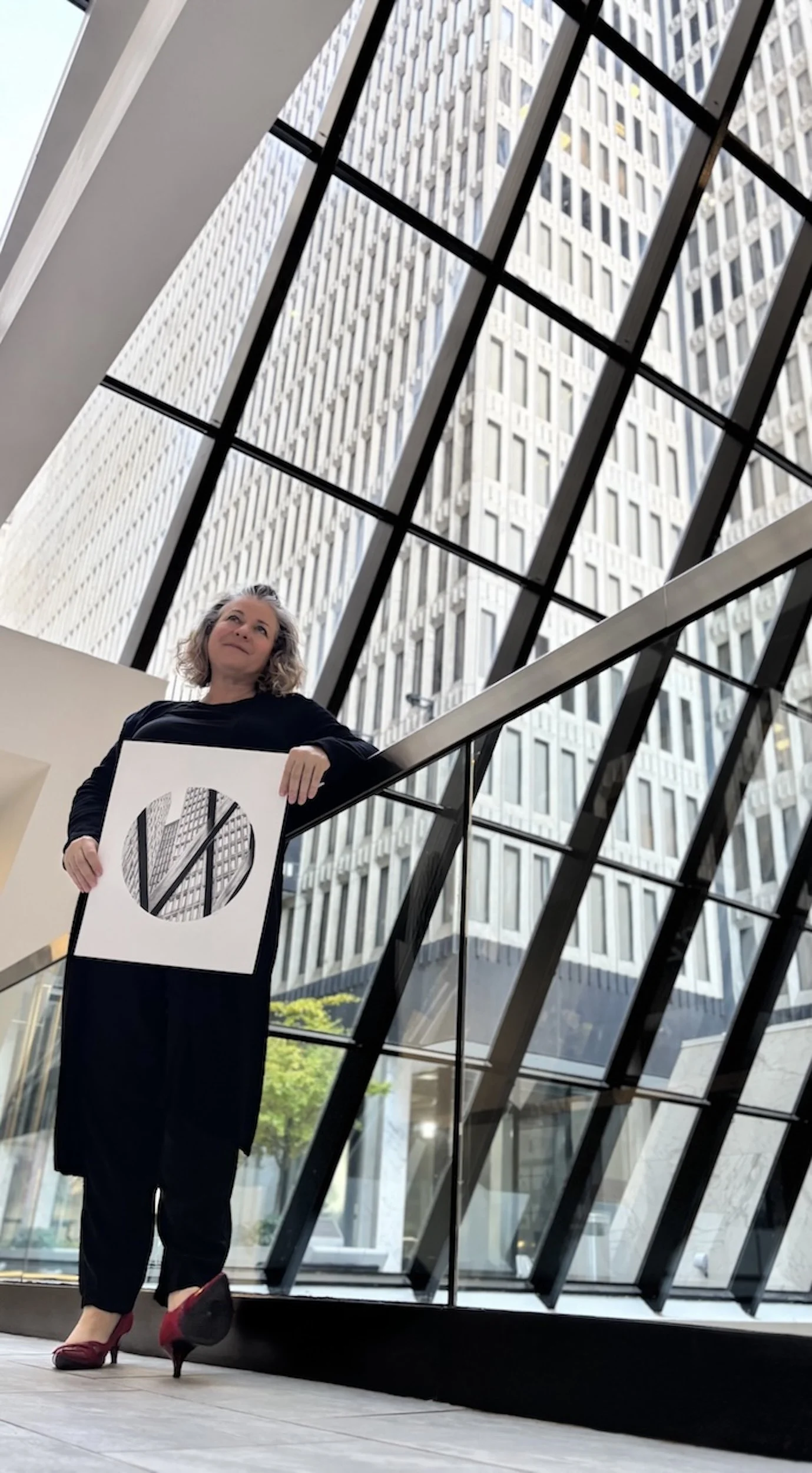 Woman with curly gray hair in black attire holding a sign with a geometric design, standing indoors in front of large glass windows with a modern building in the background.