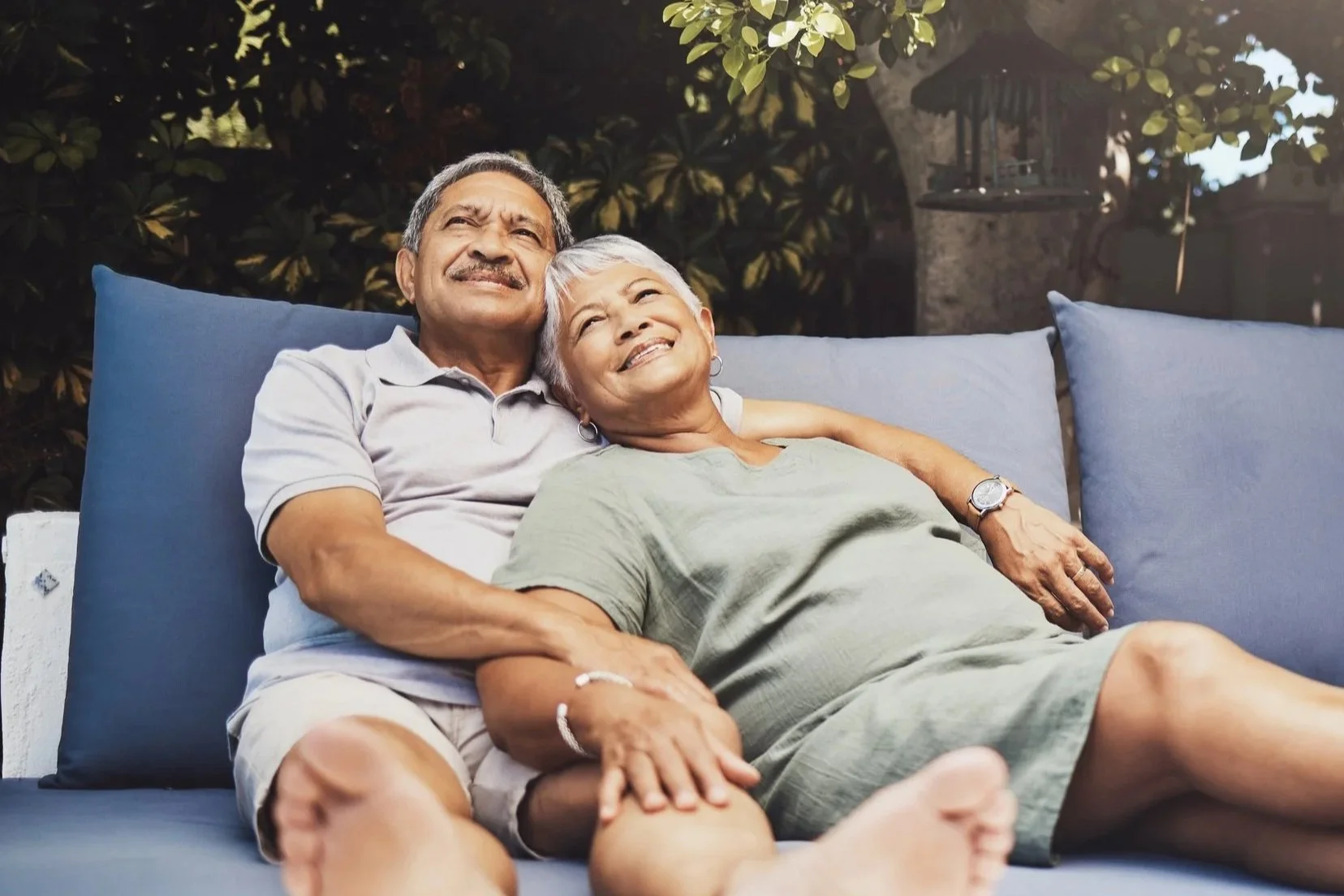 An elderly couple relaxing on outdoor sofa, smiling and looking up, surrounded by greenery.