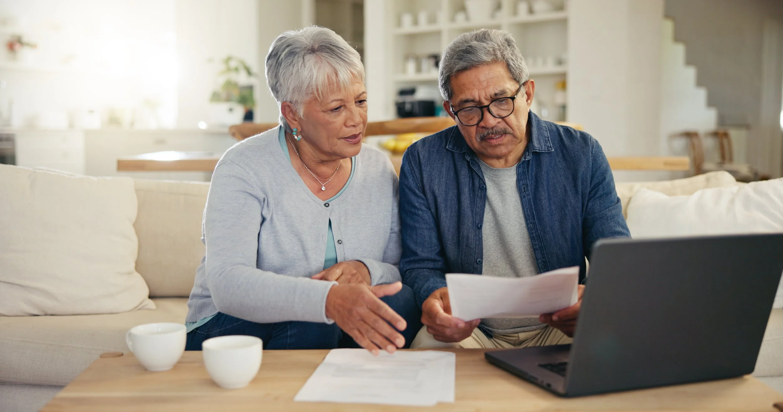 An elderly couple sitting on a couch looking at papers and a laptop together in a well-lit living room.