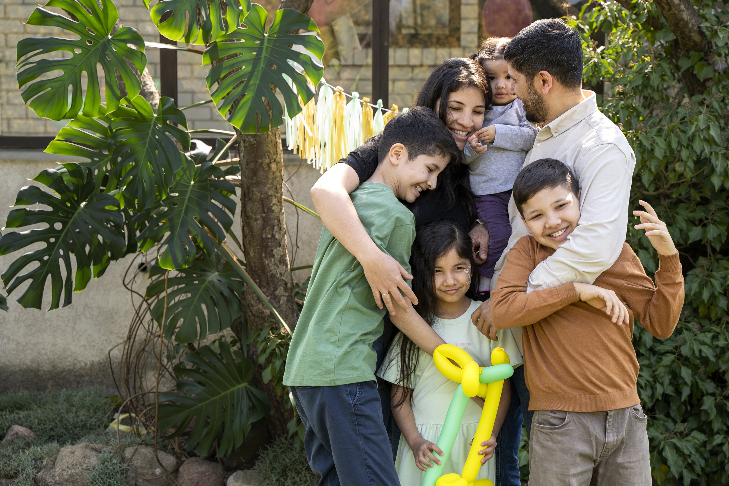 A group of people, including children and adults, hug and smile outdoors in front of large leafy green plants and a brick wall. There are some yellow balloon animals in the scene.