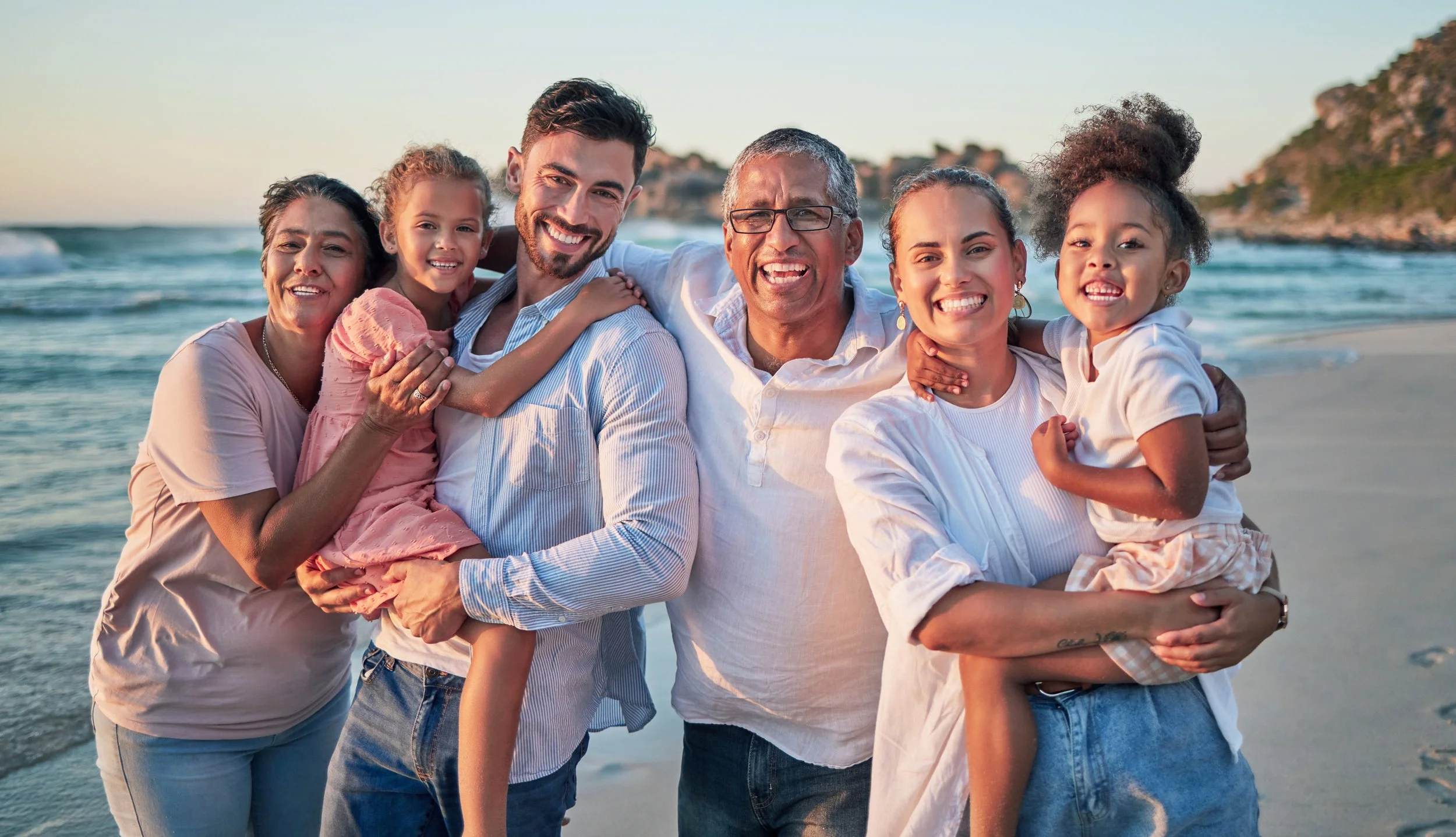 A multigenerational family of six smiling and embracing on the beach during sunset, with ocean waves and cliffs in the background.