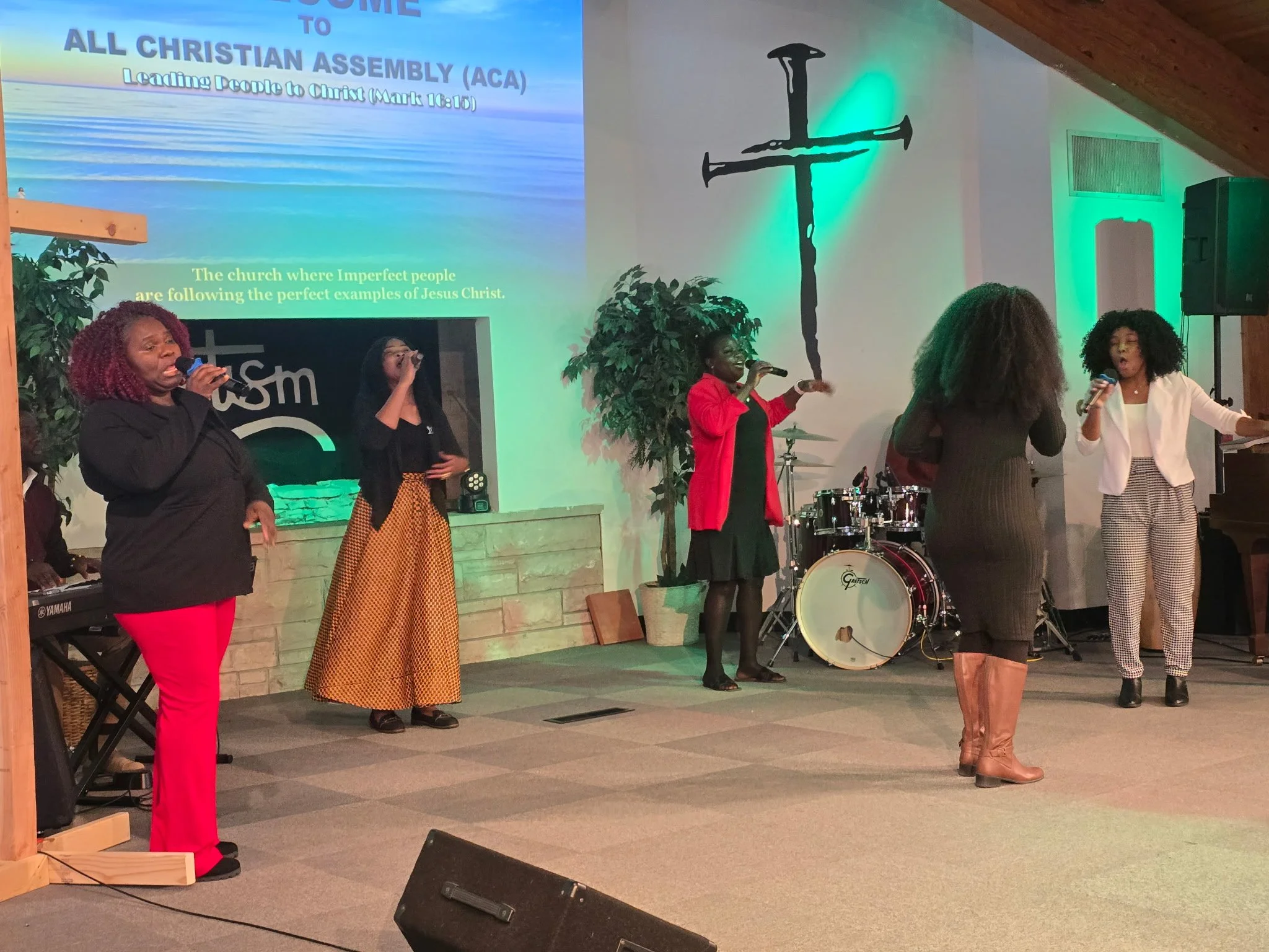 A church interior with four women singing and performing on stage, with a large screen displaying the text 'Welcome to All Christian Assembly (ACA)' and a cross painted on the wall.