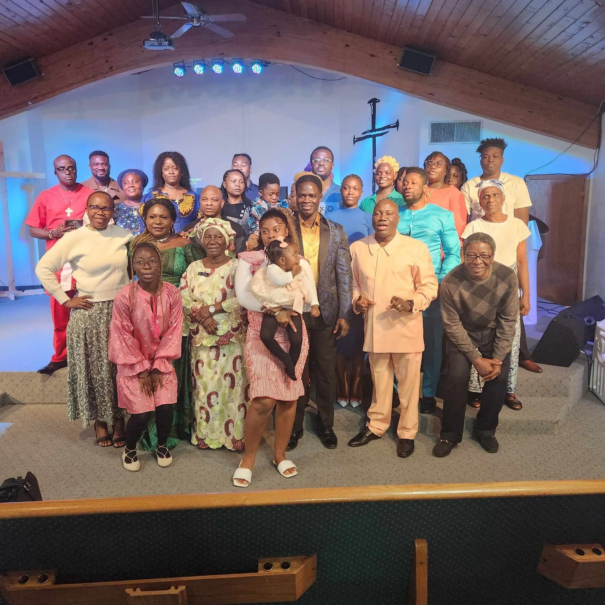 A large group of people gathered in a church with a wooden vaulted ceiling and a cross on the wall behind them. They are posing for a group photo, some smiling, with a mixture of men, women, and children of various ages.