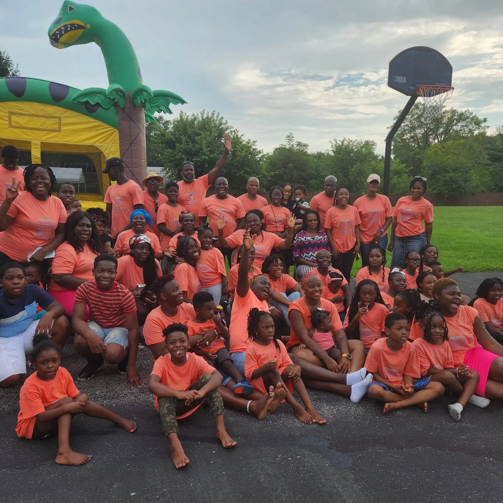 A large group of kids and adults gathered outdoors for a celebration, with many wearing orange shirts. They are smiling, waving, and sitting on the ground or standing. In the background, a colorful inflatable palm tree and a basketball hoop are visib