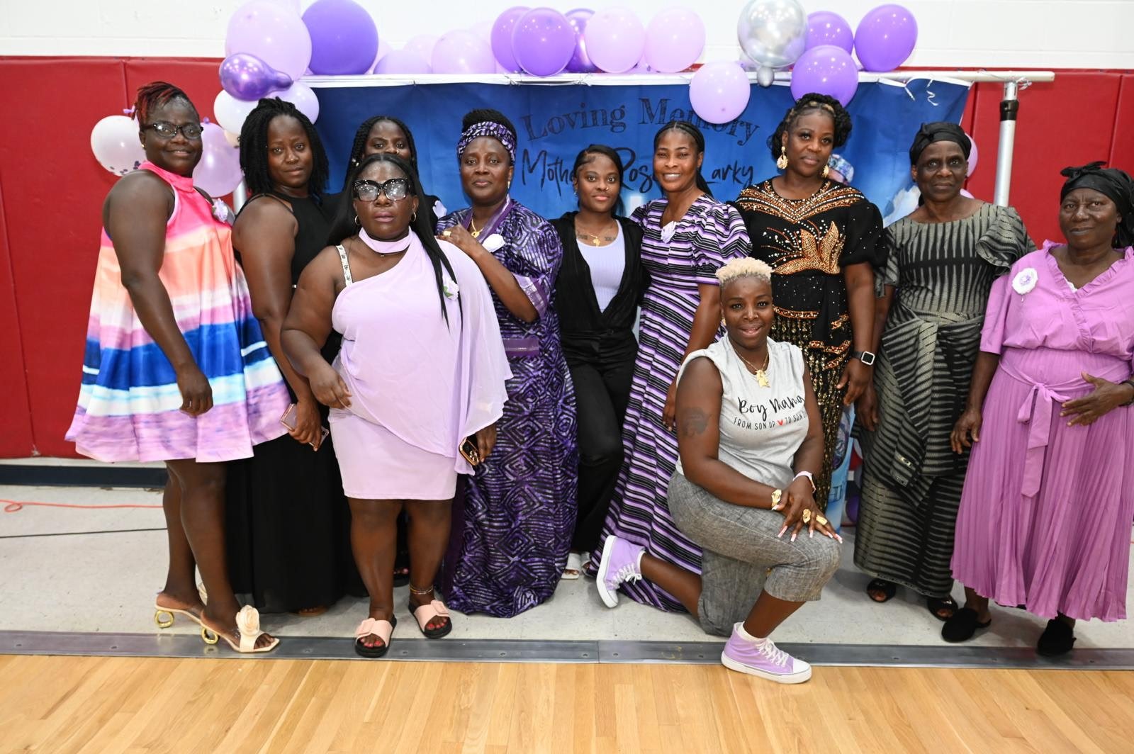 A group of women dressed in colorful traditional and modern clothing, gathered together for a photo in front of a blue backdrop decorated with purple and white balloons. The backdrop has text indicating a memorial event for 'Mother Mosby'.