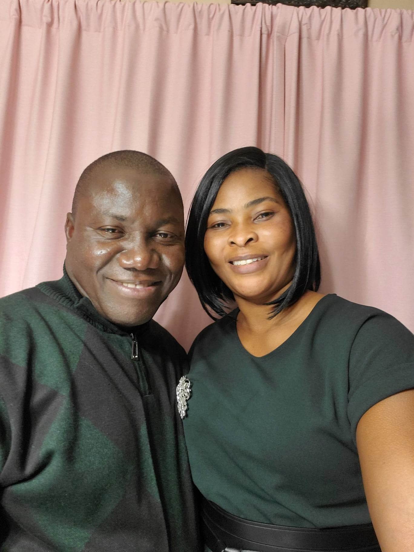 A man and woman smiling and taking a selfie in front of a pink curtain.