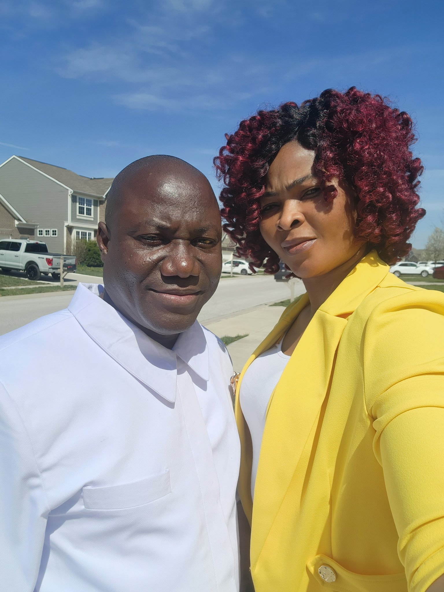 A man and a woman taking a selfie outdoors on a sunny day, with suburban houses and parked cars in the background.