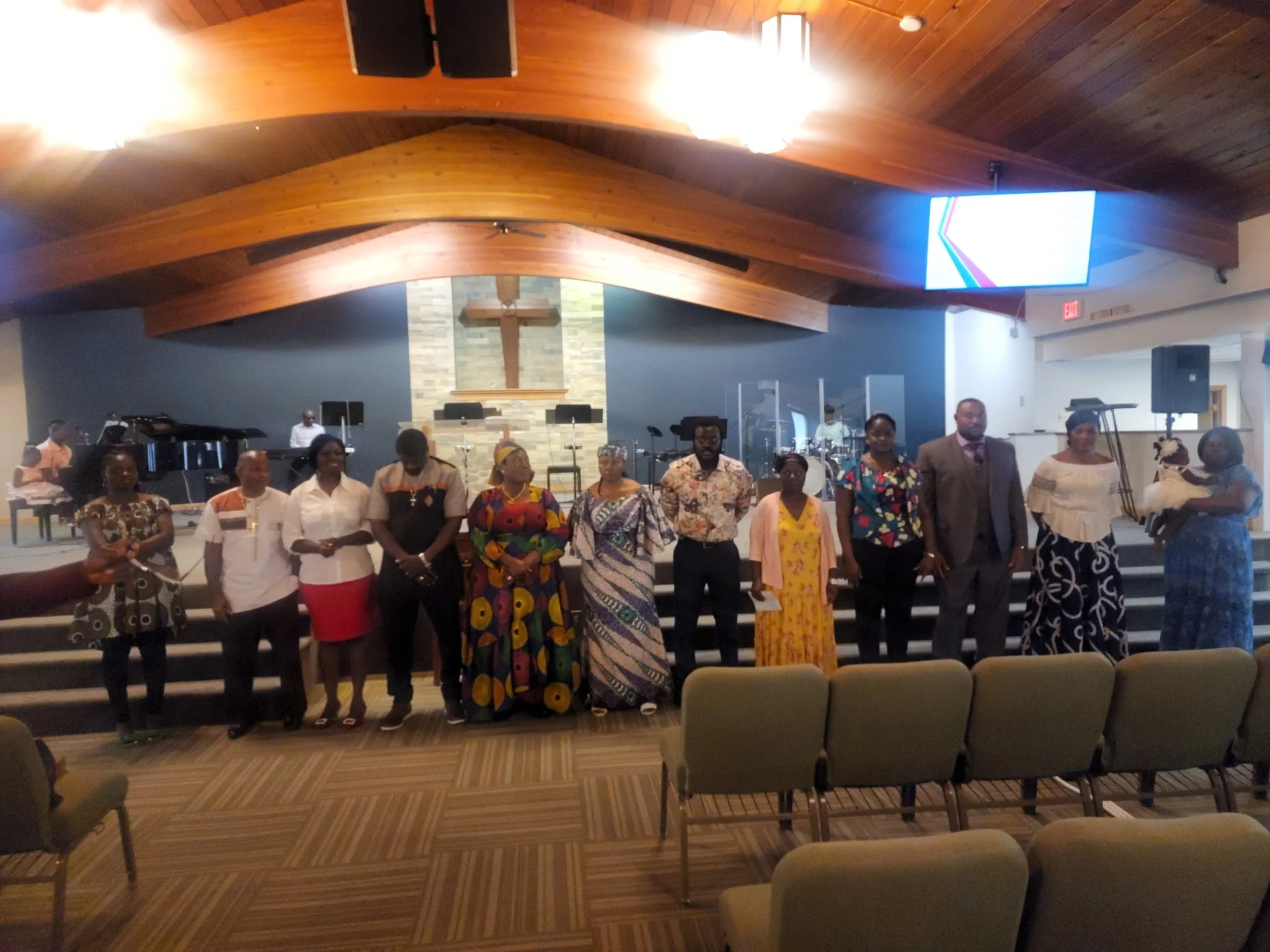 Group of people dressed in colorful clothing standing inside church or community hall with wooden ceiling, cross on the wall, and chairs arranged for an event.