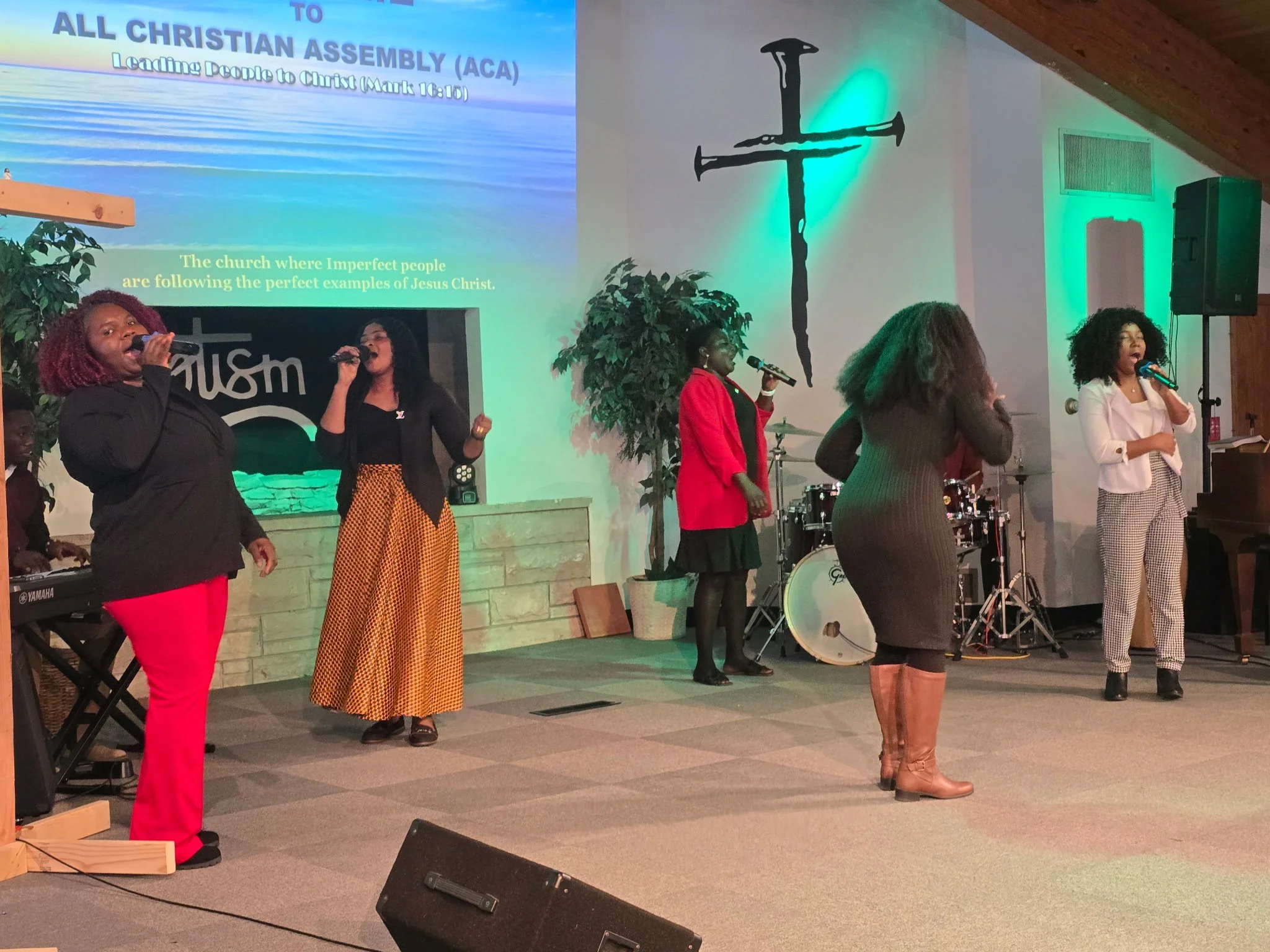 A group of women singing on stage in a church with musical instruments, a large cross on the wall, green accent lighting, and a projected Bible verse about all Christian assembly.