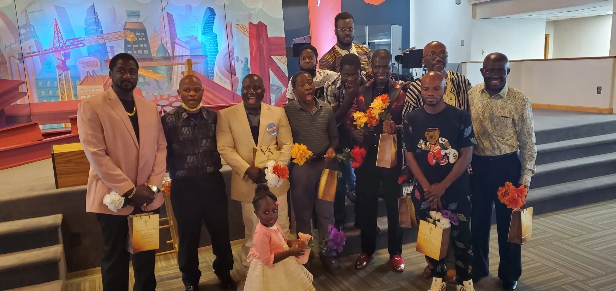 A group of eleven people, including children, gathered on a stage in a decorated indoor setting. They are holding flowers and gift bags, and some are dressed formally while others are in casual attire. The background features a colorful mural of city