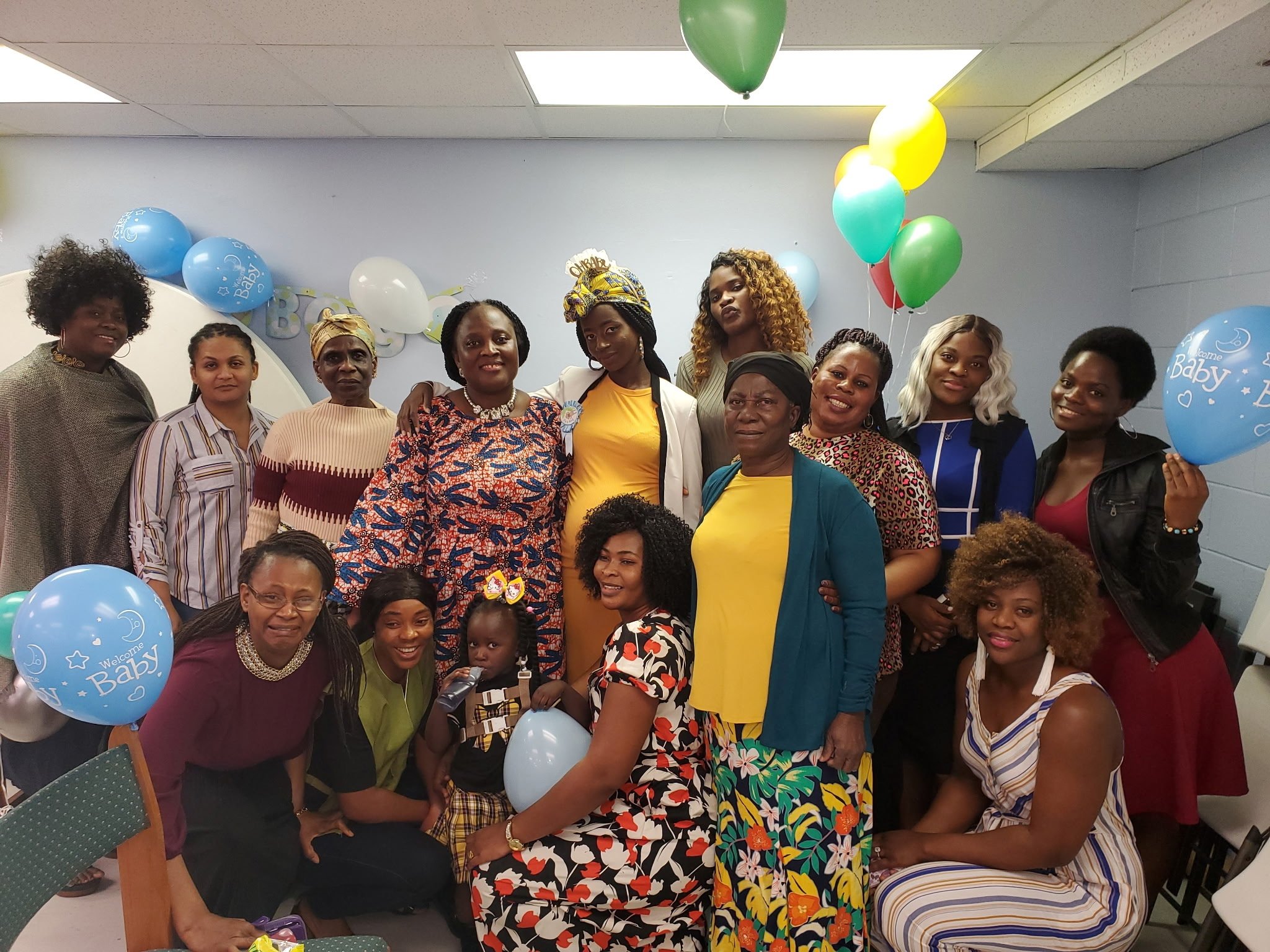 Group of people gathered at a baby shower celebration with balloons, some with 'Welcome Baby' written on them, and decorations on the wall.