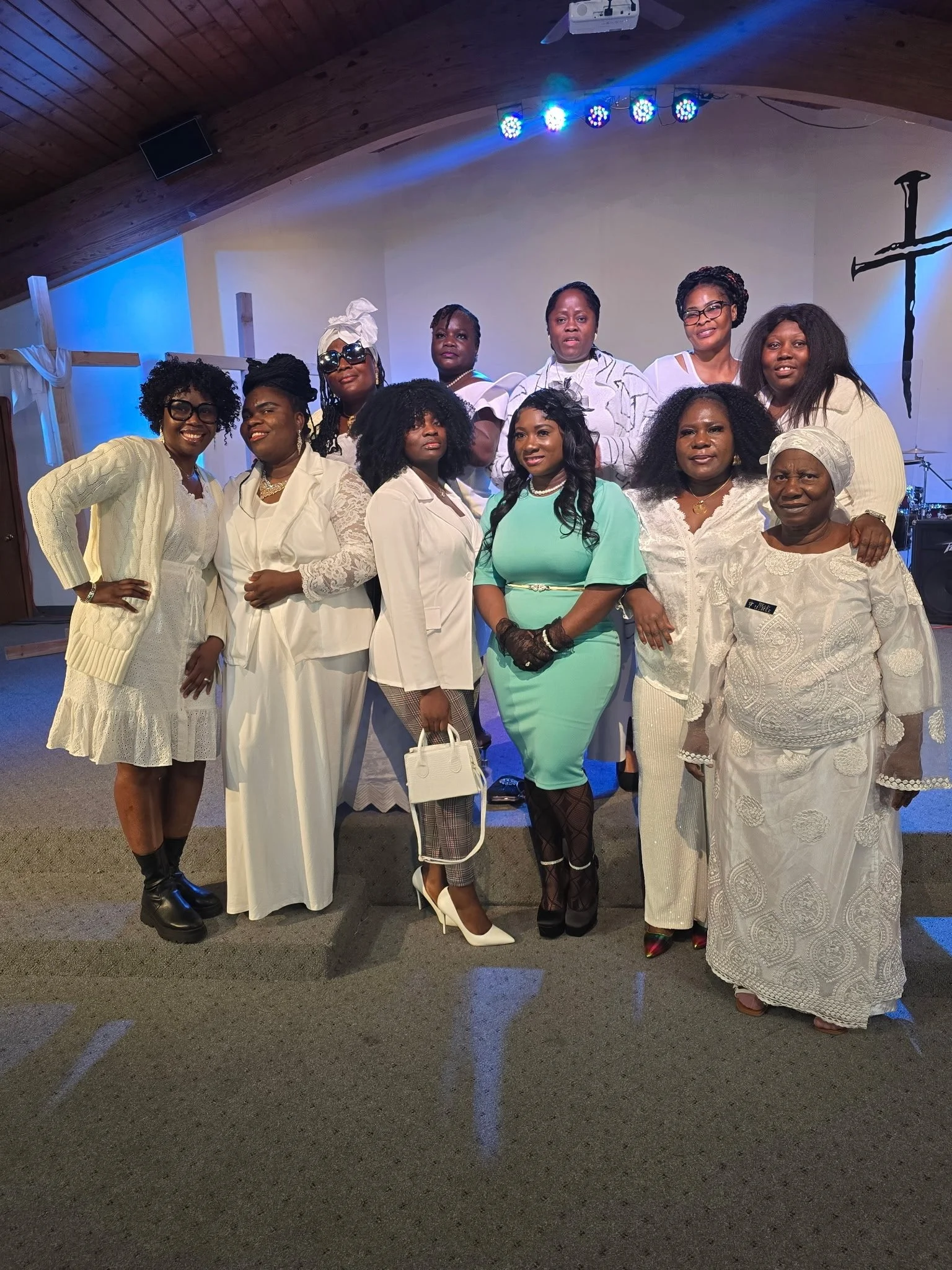 A group of twelve women dressed in white and light blue attire posing together inside a church with a cross on the wall and a stage with musical equipment in the background.