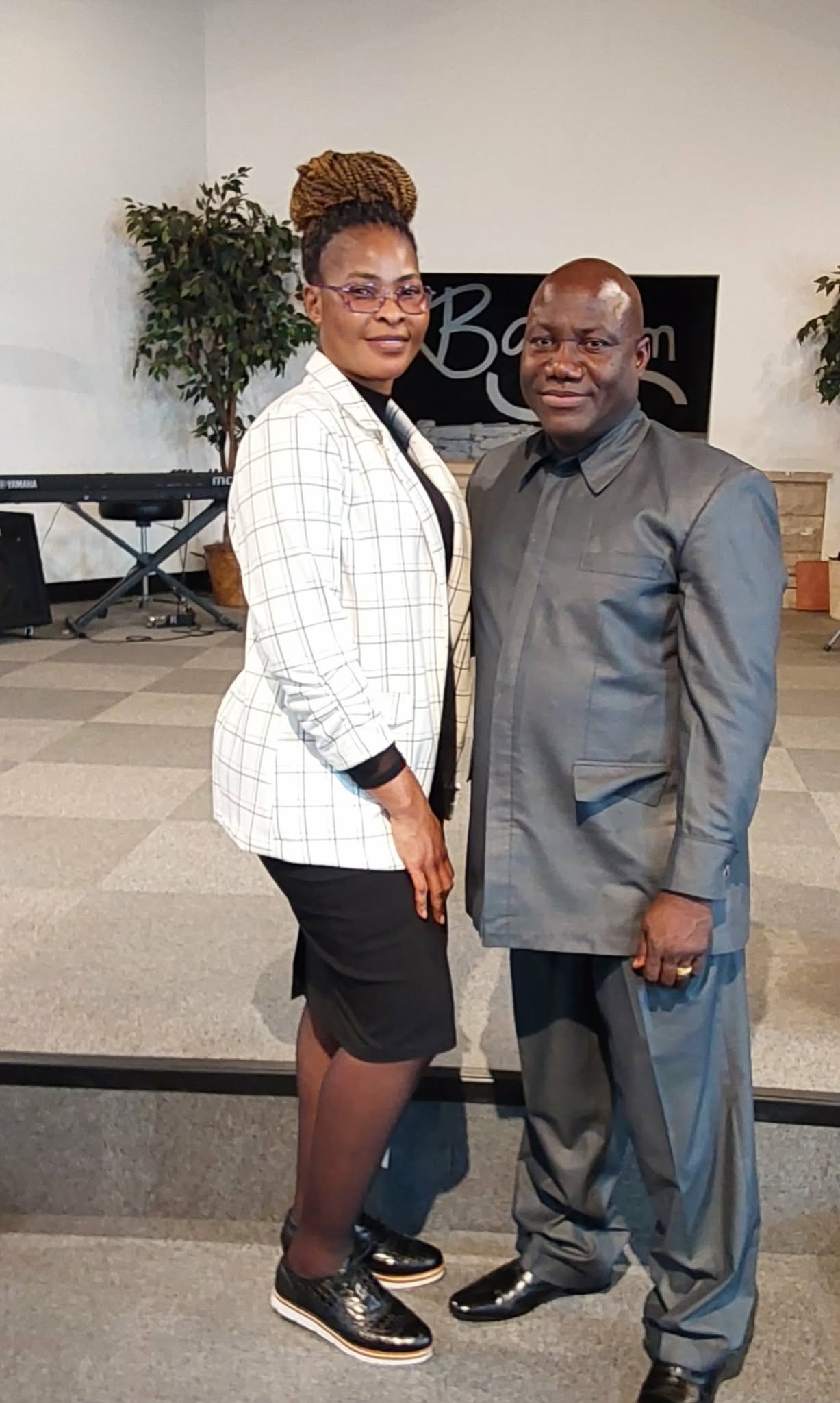 A woman and a man standing together inside a room, smiling at the camera. The woman has curly hair styled in an updo, wearing a checked blazer over a black outfit, and black sneakers. The man has a bald head, wearing a dark gray shirt and gray trouse