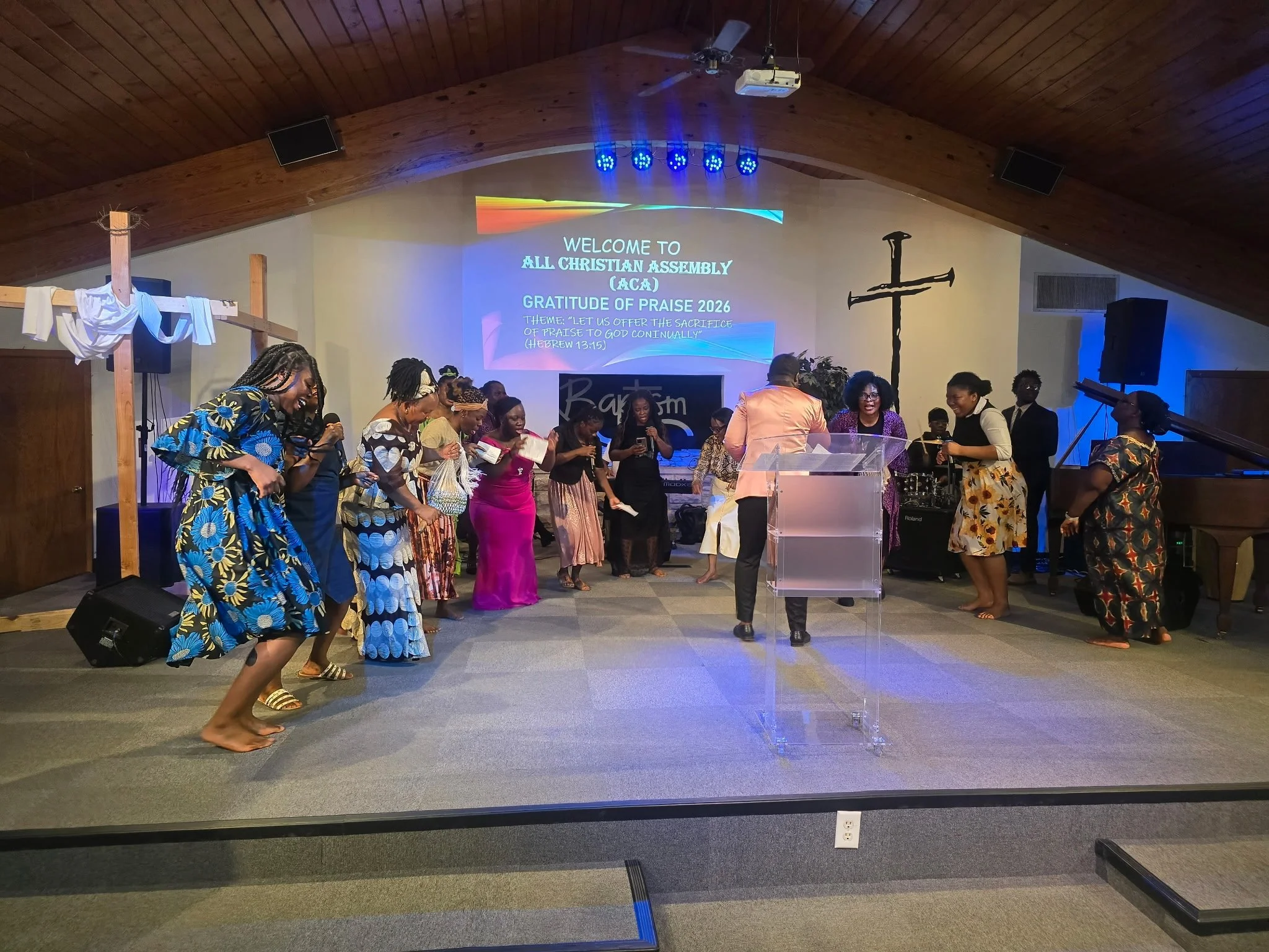 Group of diverse people, mostly women, dancing and celebrating in a church sanctuary with a large screen displaying a welcome message for a Christian assembly event.