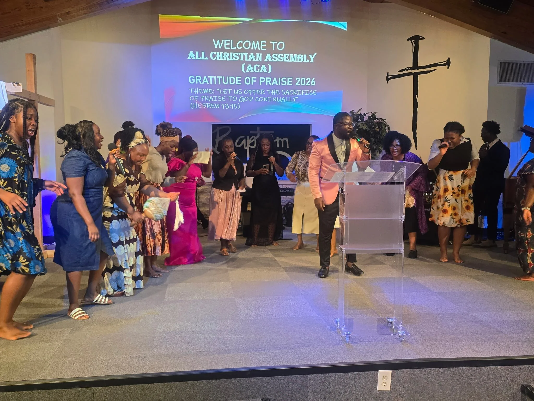 People participating in a church service or event, standing with heads bowed and hands raised, with a projection screen in the background displaying a welcome message for the All Christian Assembly Gratitude of Praise 2026 event, themed "Let us offer