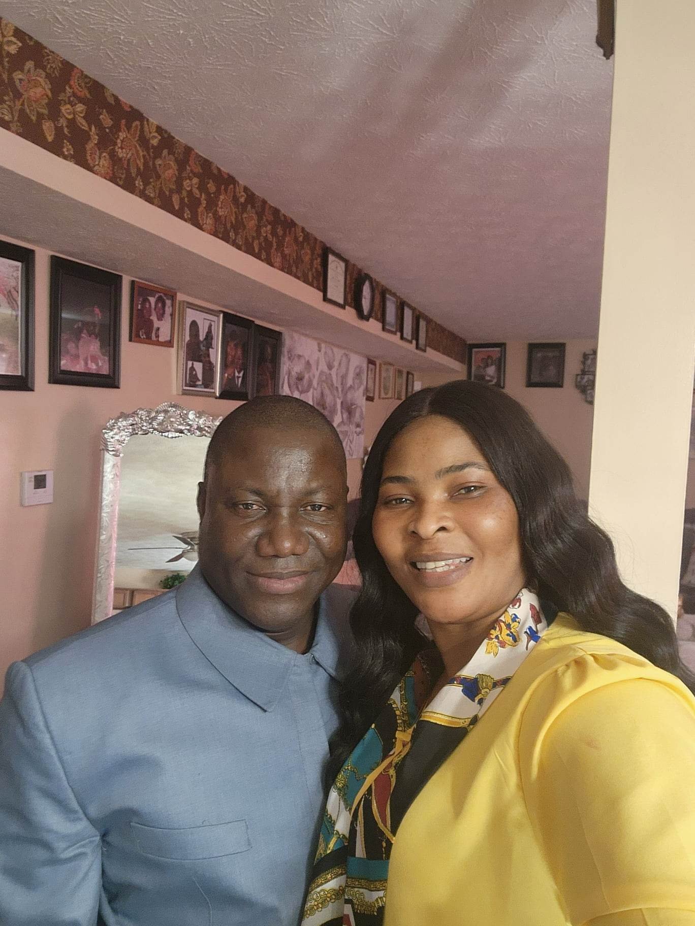 A man and woman taking a selfie in a home with framed photographs on the wall and floral wallpaper.