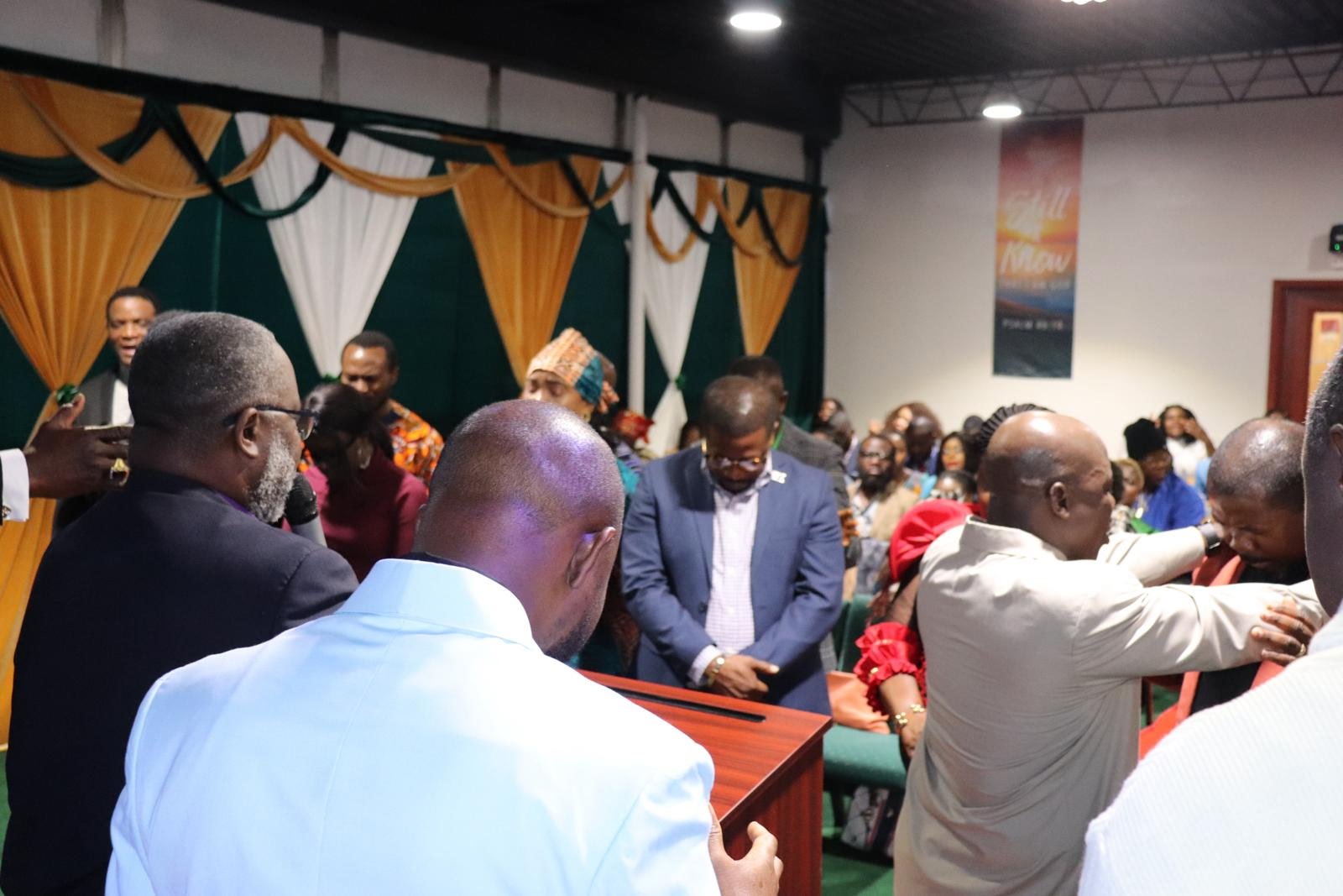 A group of people gathered in a church or conference room, some bowing their heads in prayer or reflection, with a man at a podium leading the prayer.