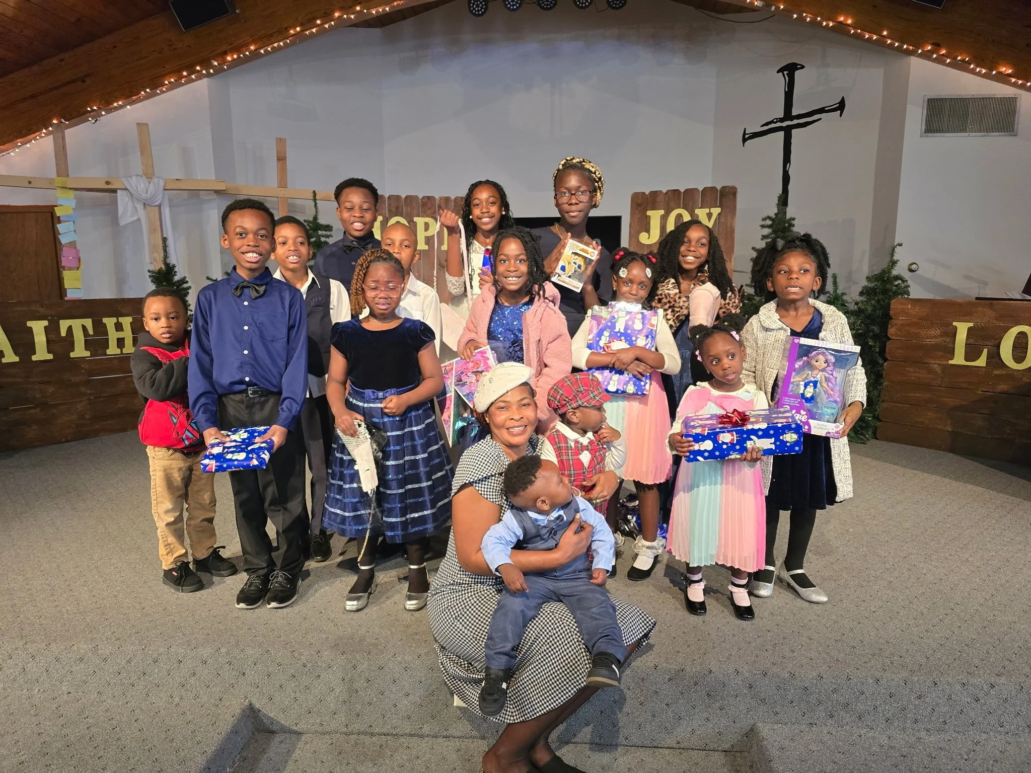 A group of children and an adult posing on a stage decorated with Christmas trees, a wooden fence with 'HOPE' and 'JOY' signs, and Christmas lights, celebrating a holiday event.