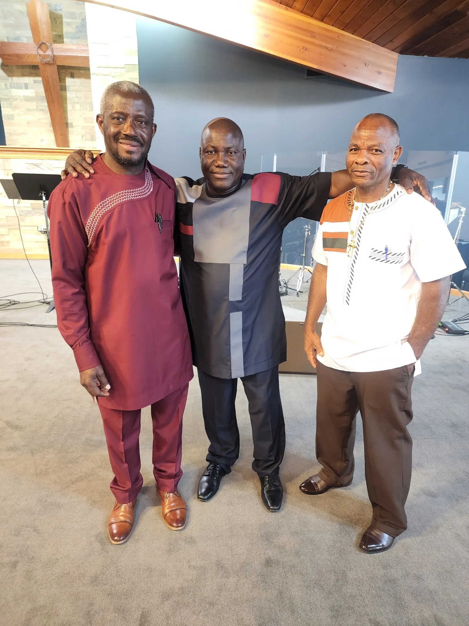 Three men standing together indoors, smiling, with musical instruments in the background.
