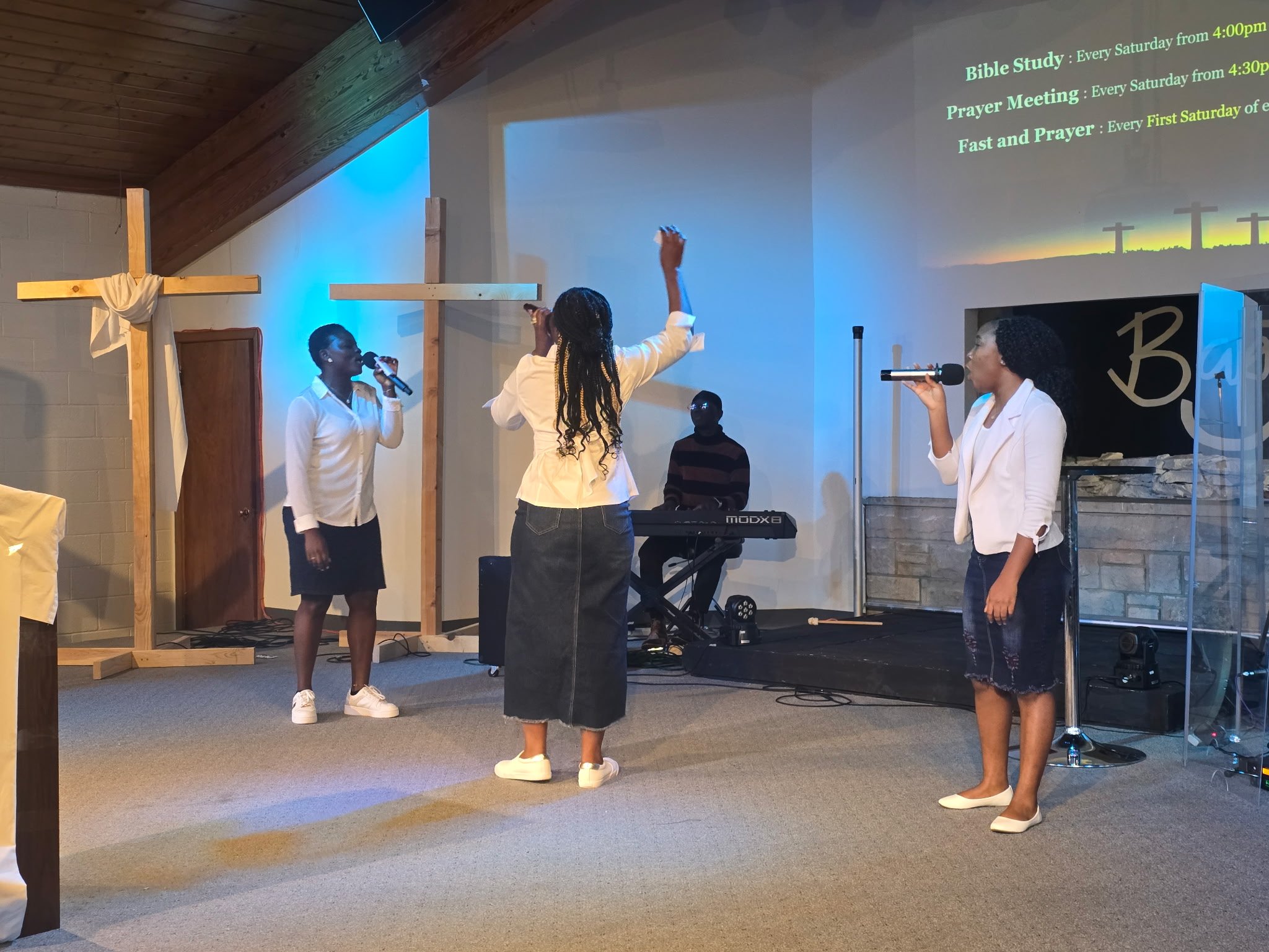 A church service or event with women singing and praying on stage, a cross decoration, and a projected church program on the wall listing activities like Bible study and prayer meetings.