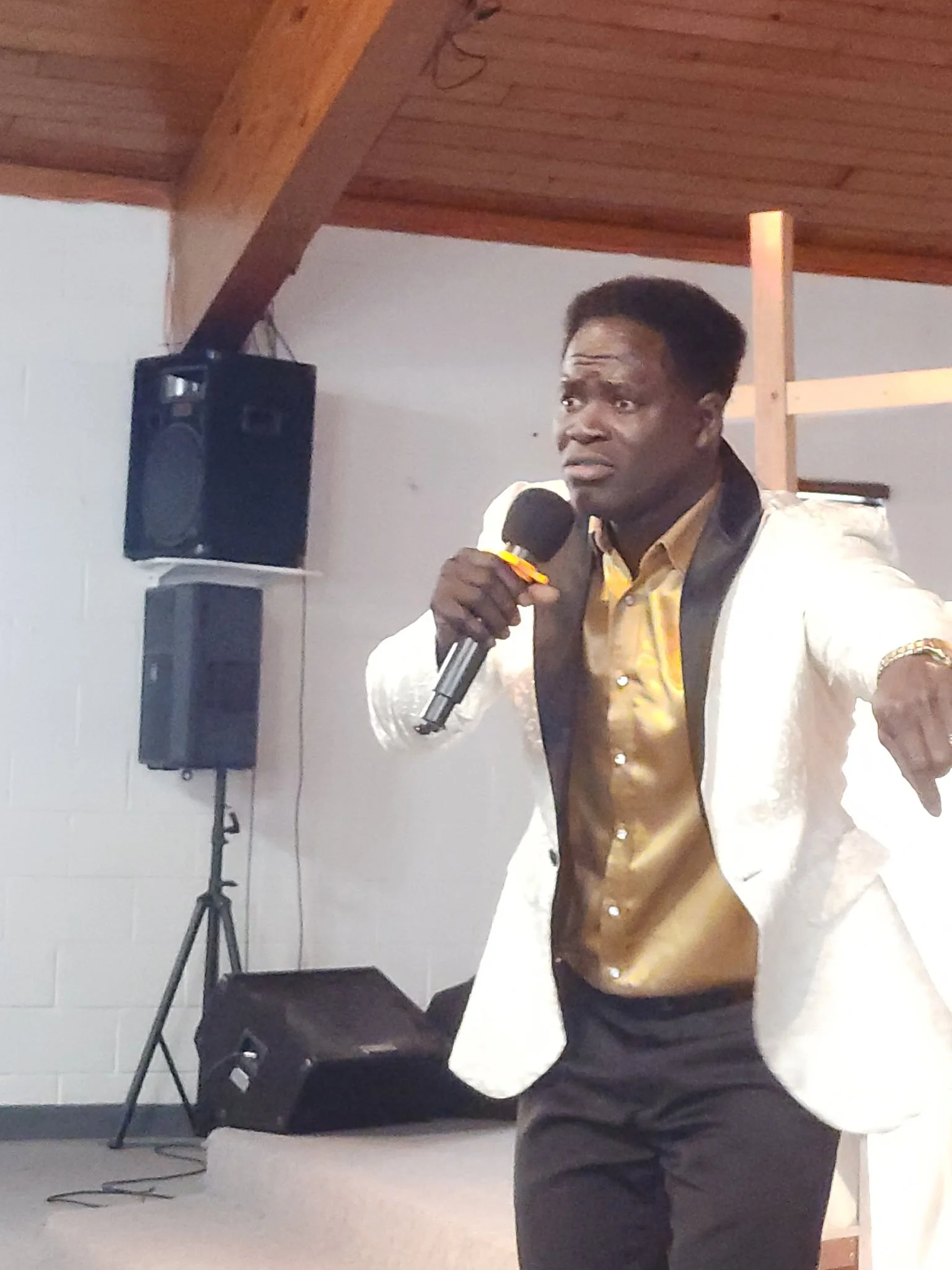 A man dressed in a shiny gold vest and a white jacket, holding a microphone, speaking or singing in a room with speakers and a wooden ceiling.
