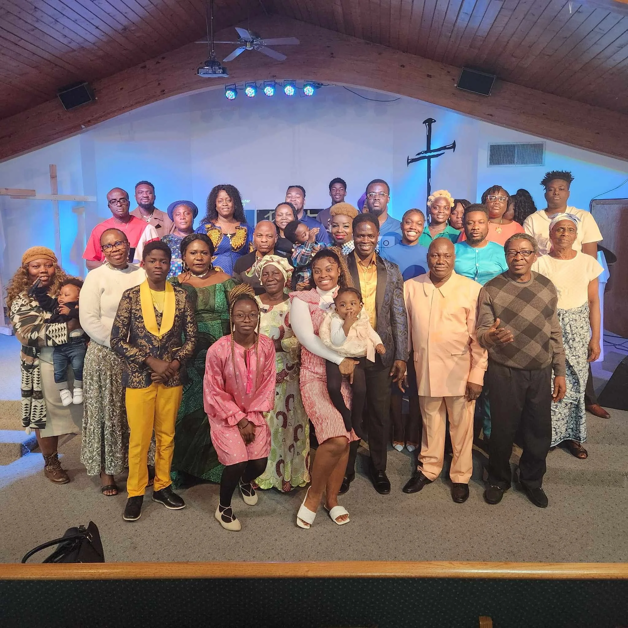 Group of diverse people gathered in a church or community hall, posing for a group photo. The wooden ceiling with a fan and lights, and a cross on the wall in the background, suggest a church setting.
