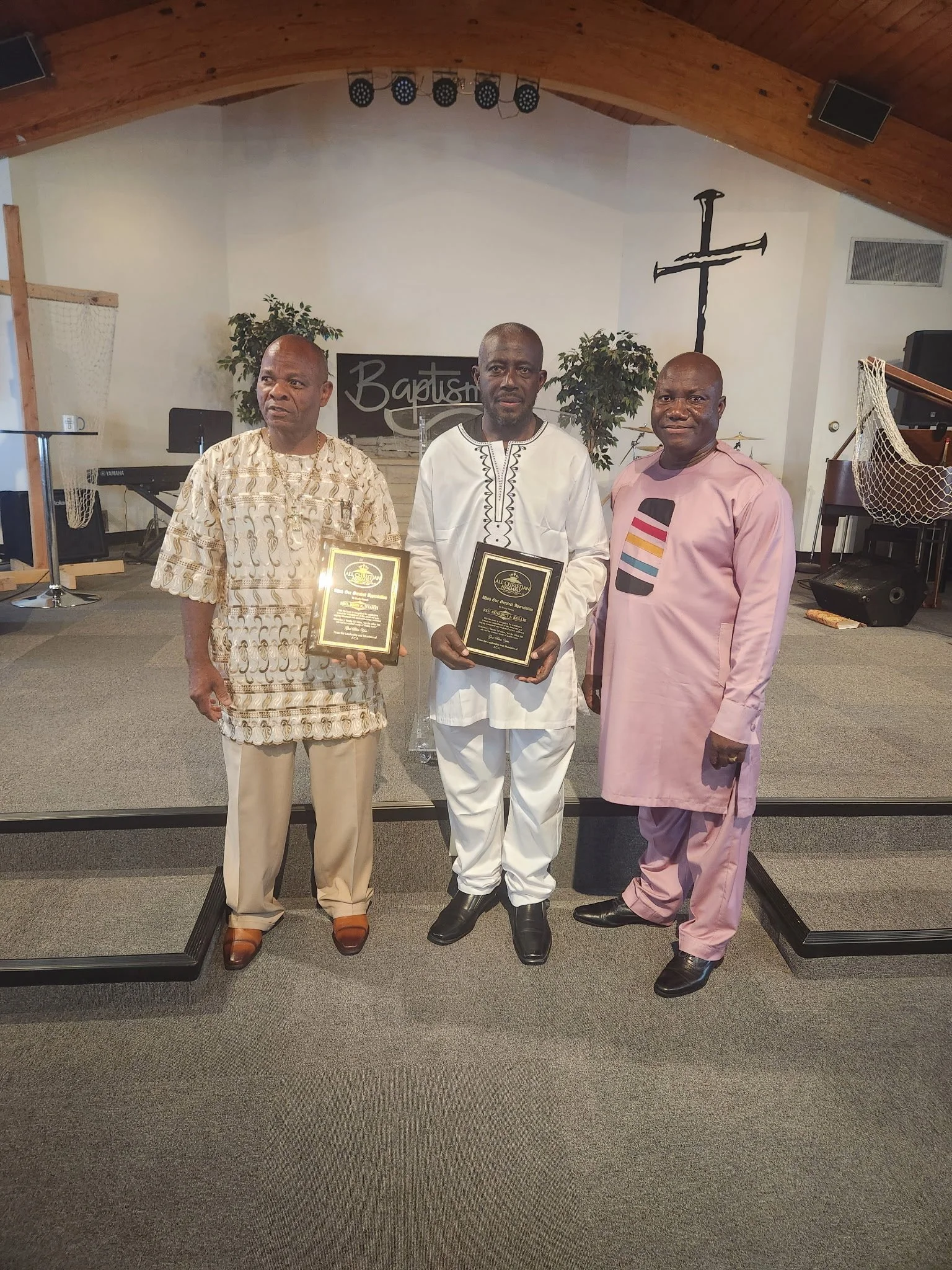 Three men standing inside a church, holding plaques, with a cross on the wall behind them and musical equipment around.
