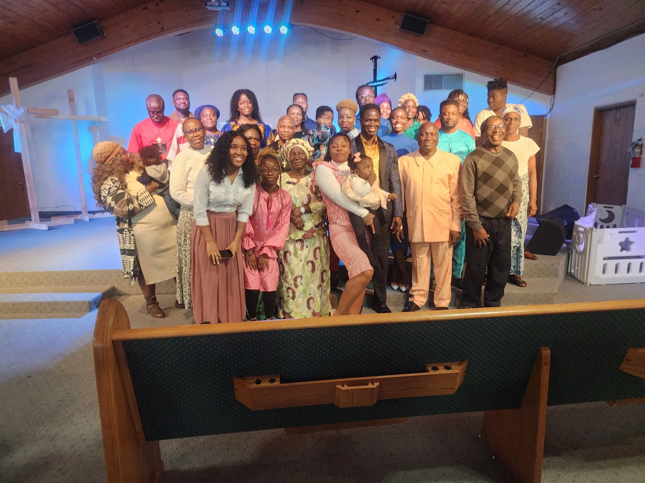 Group of people gathered together on stage for a photo in a church or community hall, with a wooden ceiling and a large cross on the wall behind them.