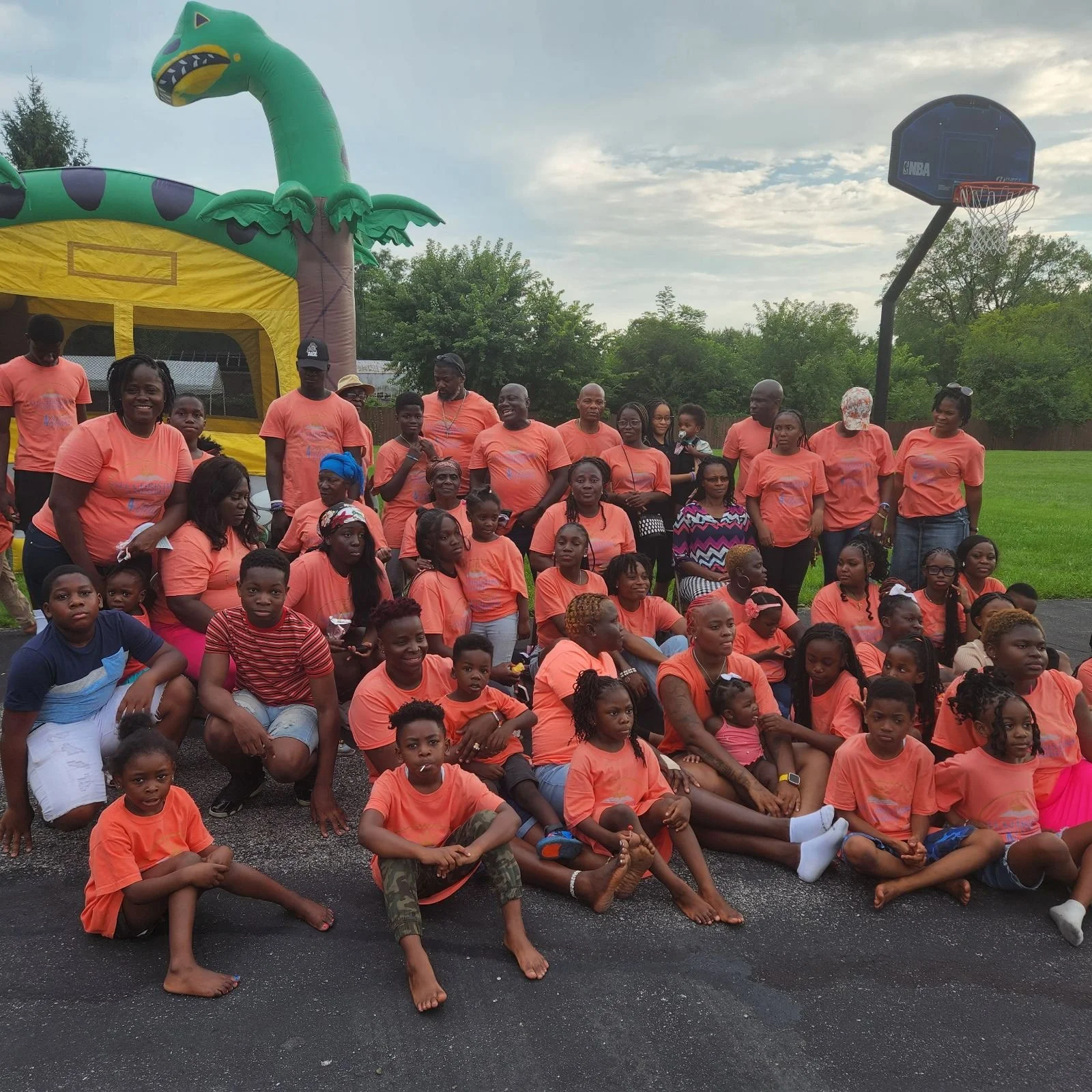 A large group of people, including adults and children, gathered outdoors on an overcast day, some sitting on the ground and others standing, with a colorful inflatable palm tree and a basketball hoop in the background.