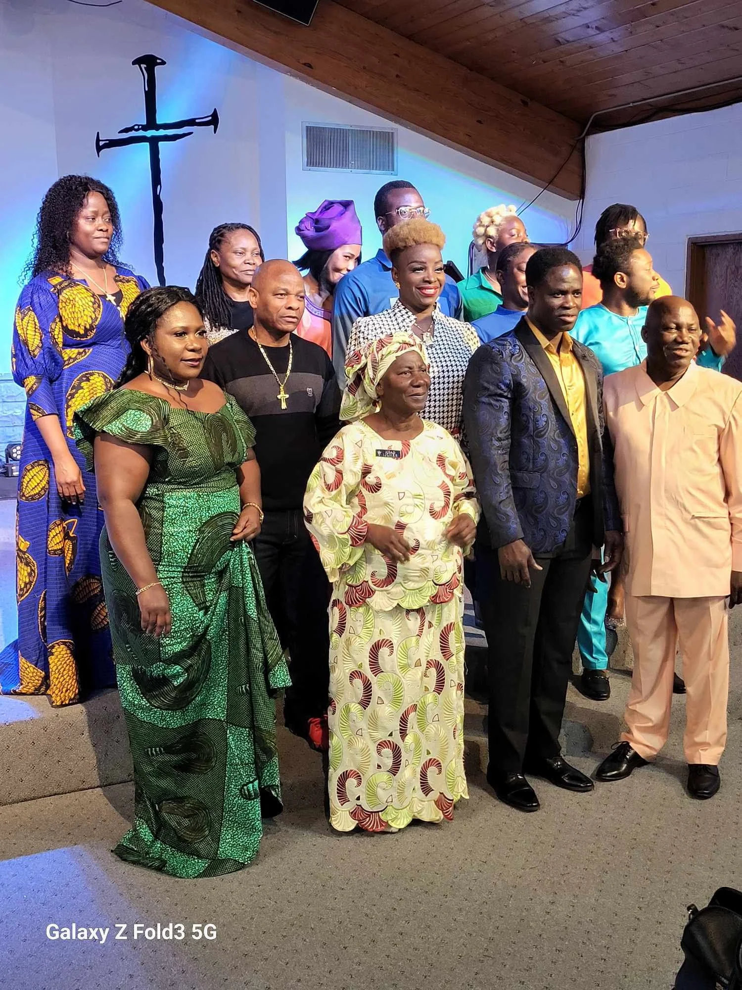 Group of people standing inside a church, some wearing colorful traditional African attire, with a cross on the wall behind them and a sign that says 'Galaxy Z Fold3 5G' at the bottom left.