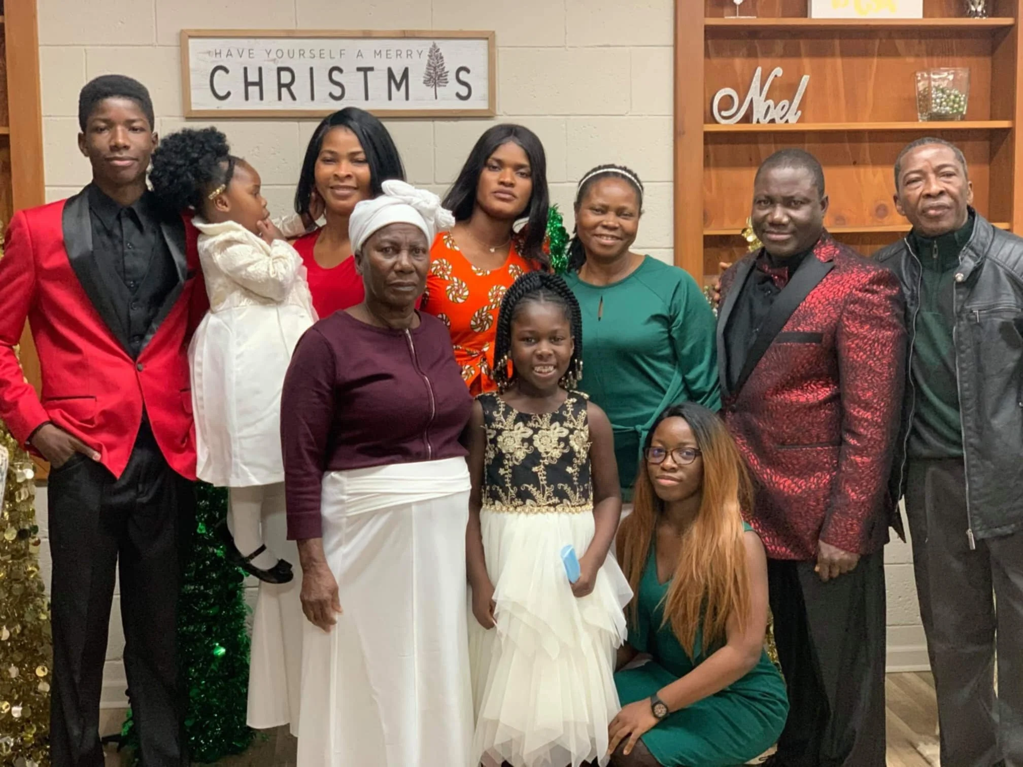 Group of people posing in front of a Christmas-themed background, including a sign that says "Have Yourself a Merry Christmas" and Christmas decorations like ornaments and a tree.