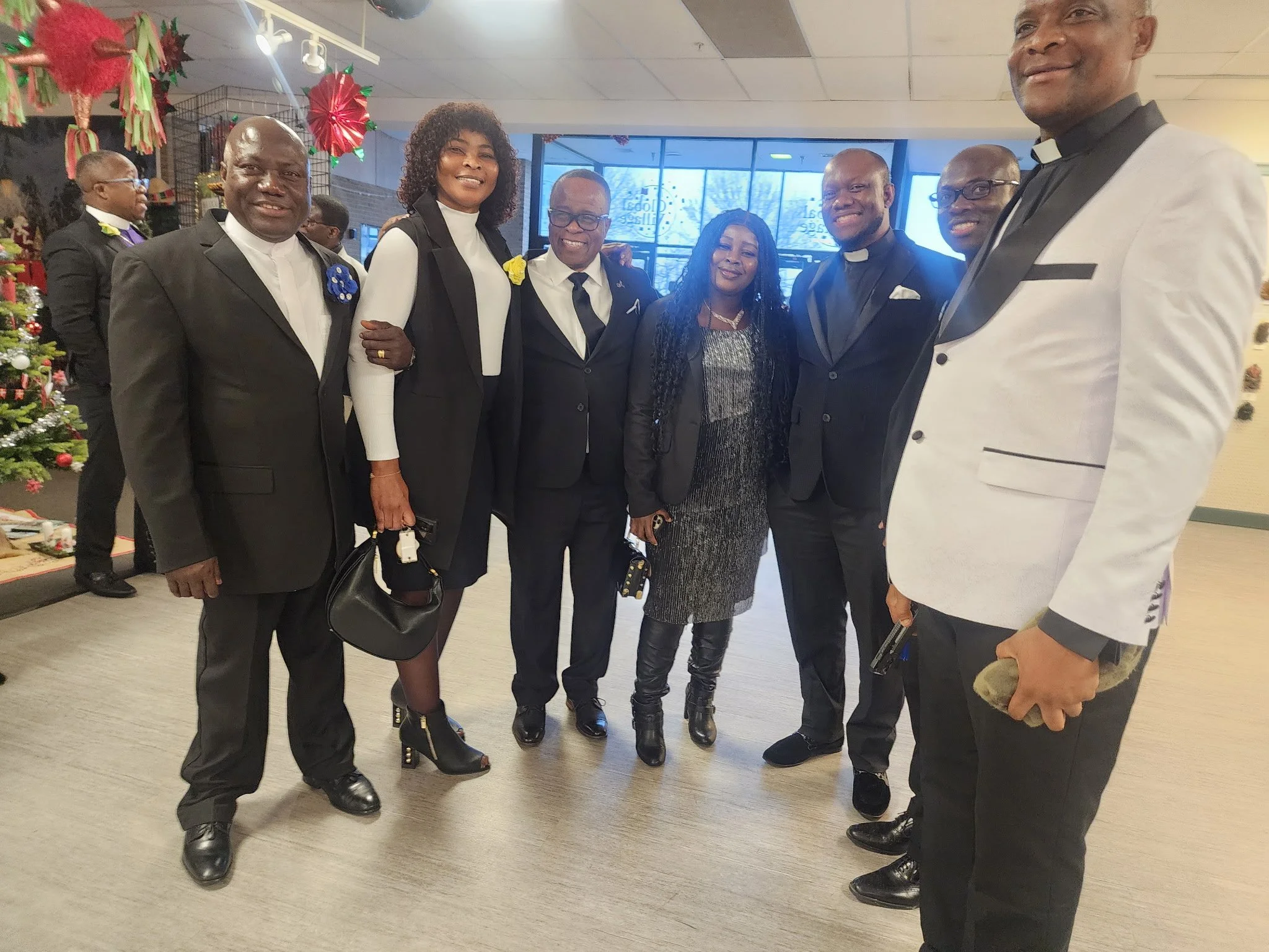 A group of seven people, including three women and four men, dressed in formal attire, standing together in an indoor setting decorated with Christmas ornaments, smiling at the camera.