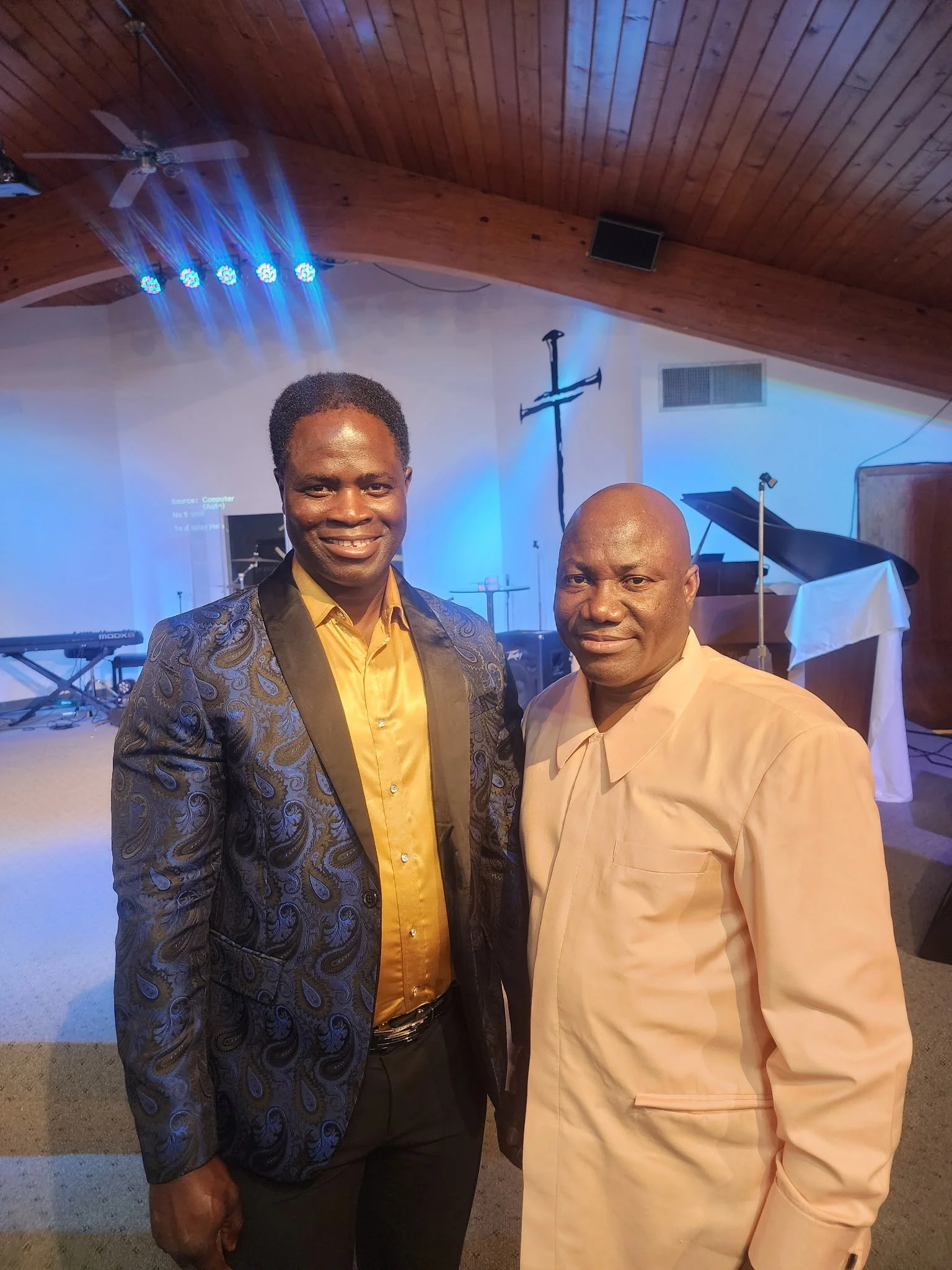 Two men posing for a photo inside a church, with a wooden ceiling, a cross, a piano, and stage lights in the background.