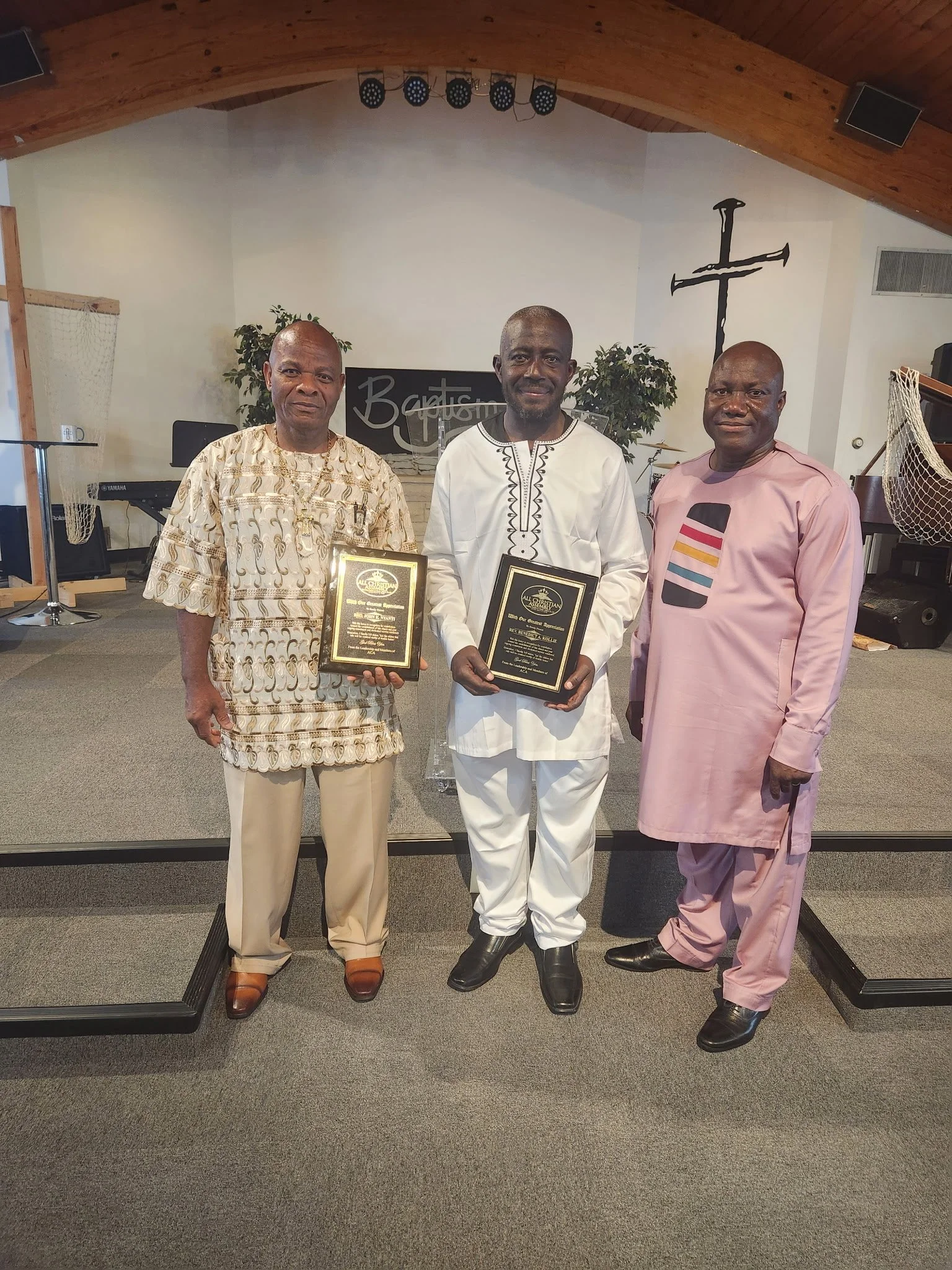 Three men standing inside a church, with two holding plaques, all dressed in traditional or formal clothing. The background features a cross on the wall, a music stand, and musical instruments.