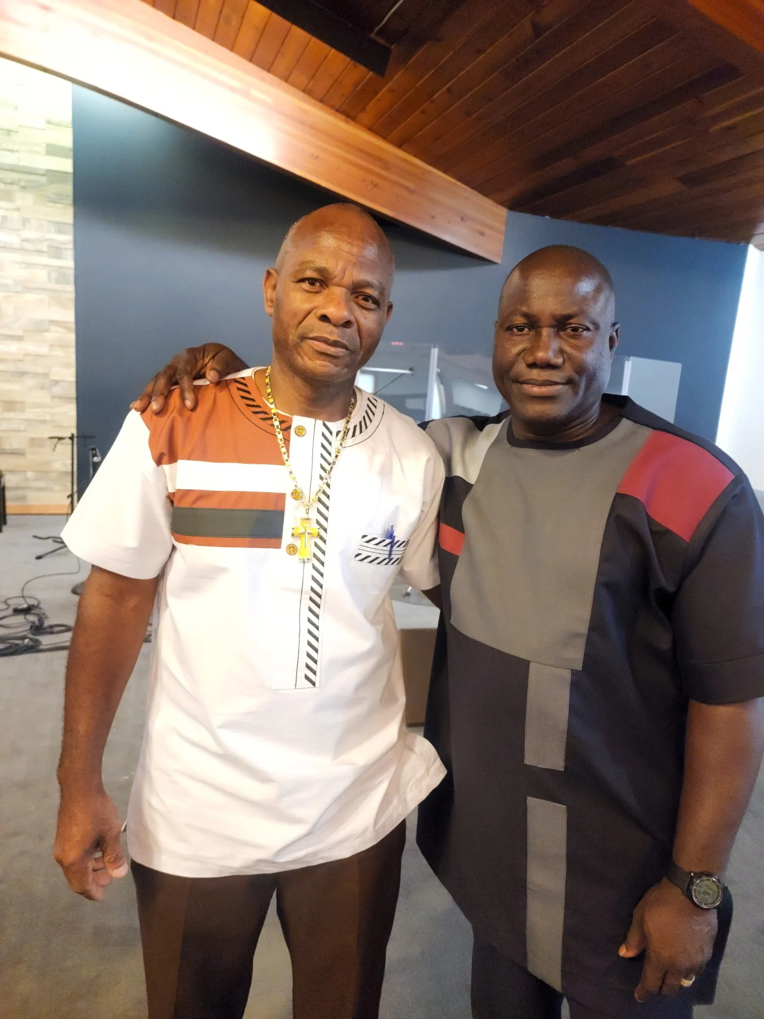 Two men standing side by side in an indoor setting, smiling slightly at the camera. One man is wearing a white and brown patterned shirt with a gold chain and a cross pendant, and the other is in a gray and black t-shirt with red accents and a wristw