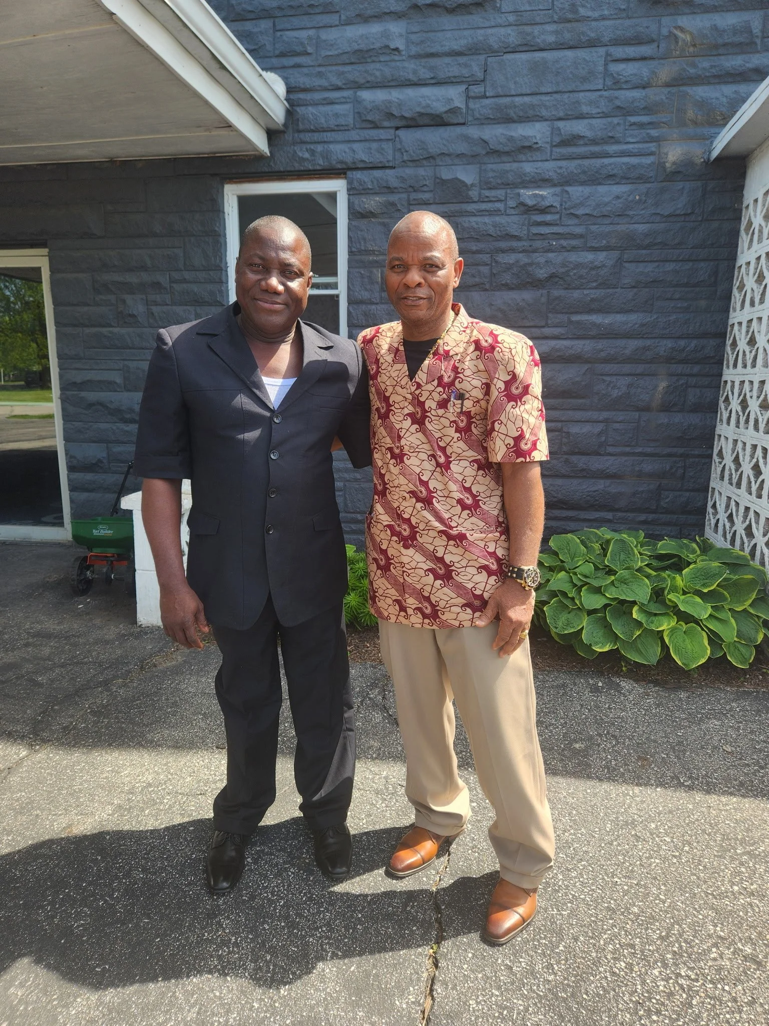 Two men posing outdoors in front of a stone wall, one in a black suit and the other in a patterned shirt and beige pants, with plants and a window in the background.
