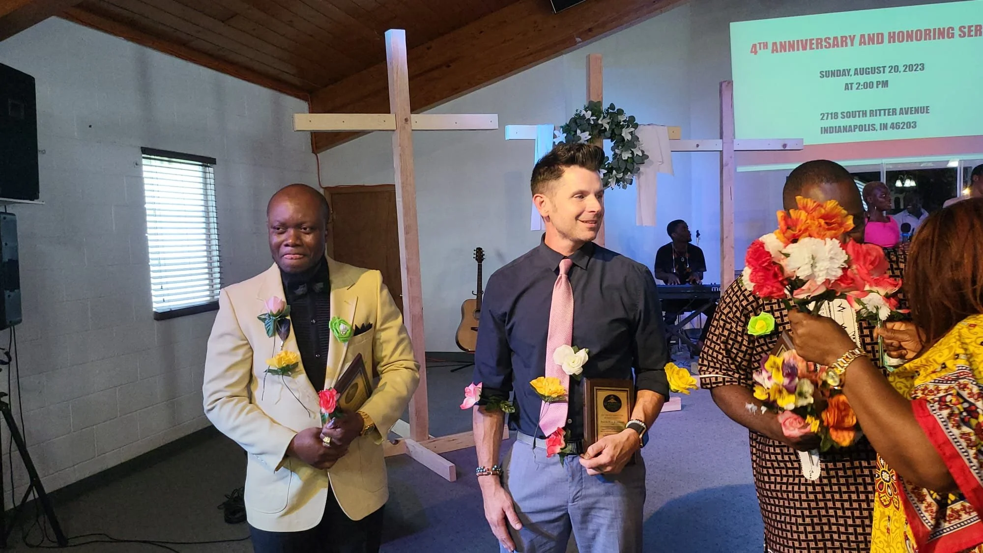 People attending an outdoor celebration with a wooden cross decorated with flowers, some holding plaques, and a screen displaying event details in the background.