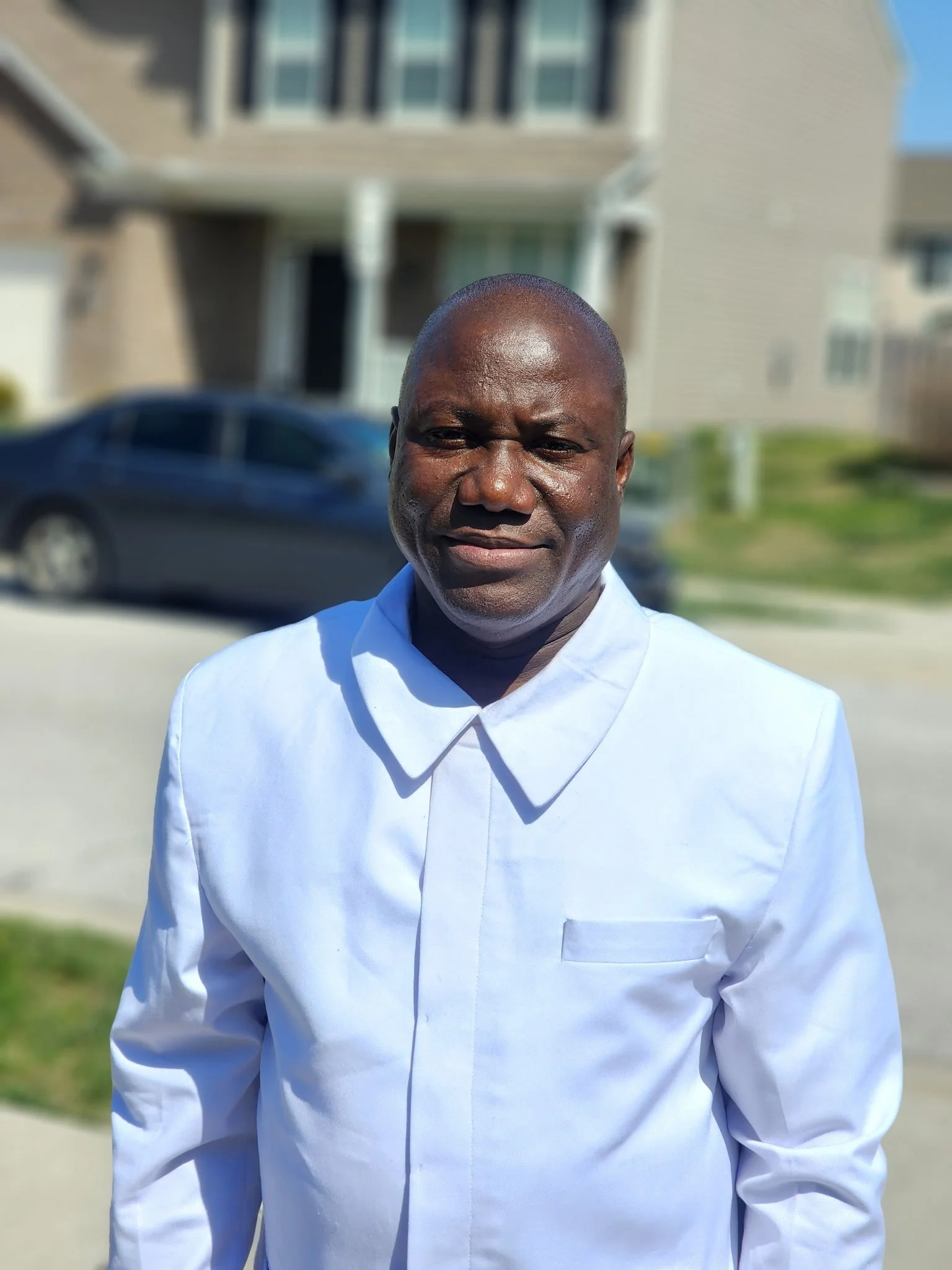 A man with dark skin, bald head, wearing a white dress shirt, standing outdoors in front of a residential building and a parked car, smiling slightly.