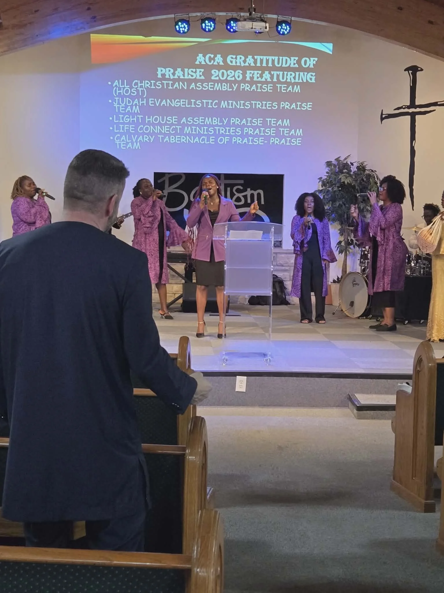 Church interior with a group of women singing at a service. A large screen displays a message of gratitude and praise for various praise teams. The church has wooden pews and a cross painted on the wall to the right.