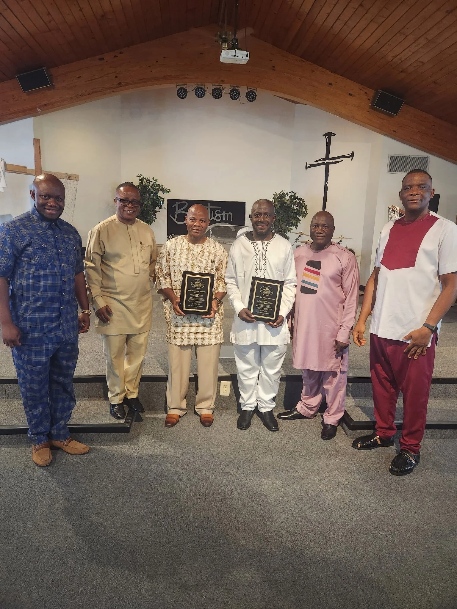 Six men standing in a church, two of them holding plaques, with a cross on the wall behind them.
