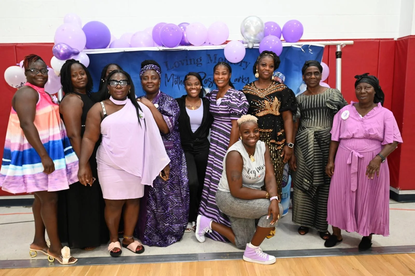 A group of women dressed in colorful dresses and traditional African attire, posing for a photo in front of a backdrop decorated with purple and white balloons at a social event.