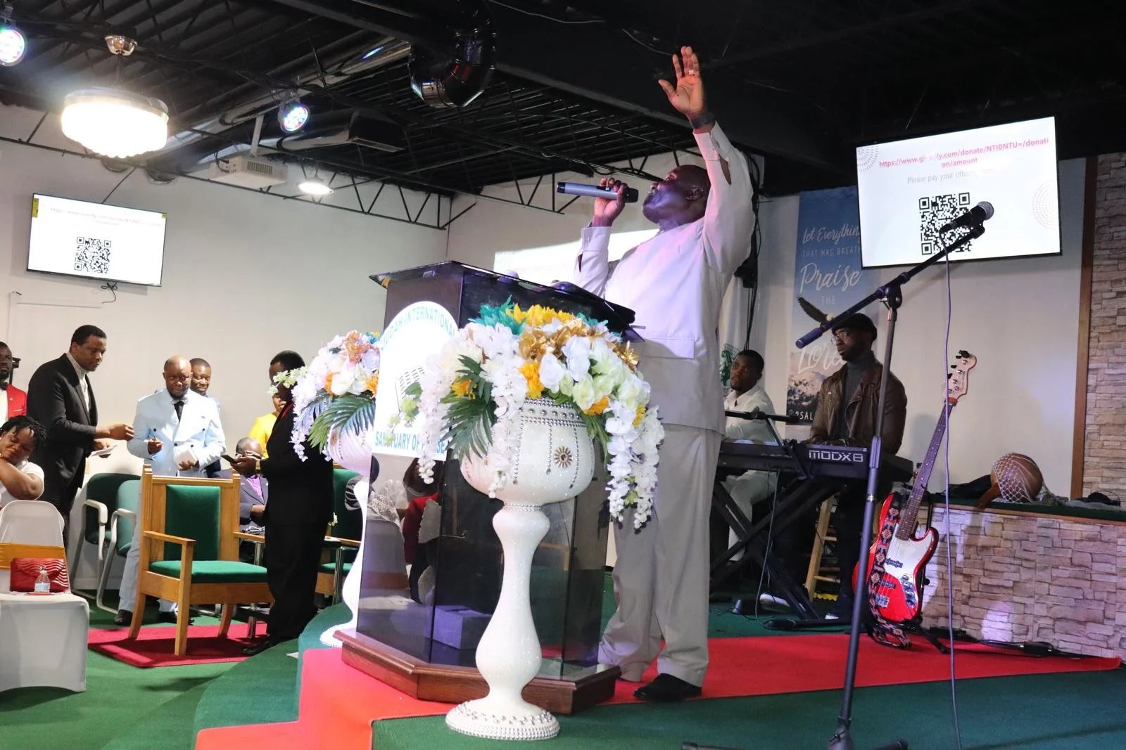 A church service or religious gathering with a man in light-colored attire speaking at a pulpit decorated with a large floral arrangement. Several people are present, some standing and some seated, with musical instruments like a keyboard and a guita