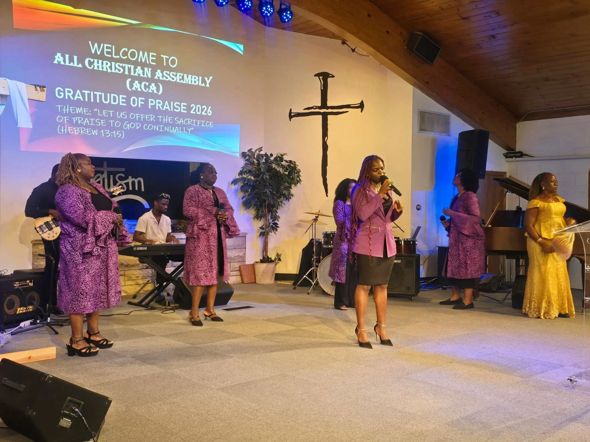 A group of women and a man worshipping and singing on stage during a church service. The women are dressed in colorful dresses, two in purple and one in gold. The man is playing a keyboard, and a female singer is holding a microphone. The background 