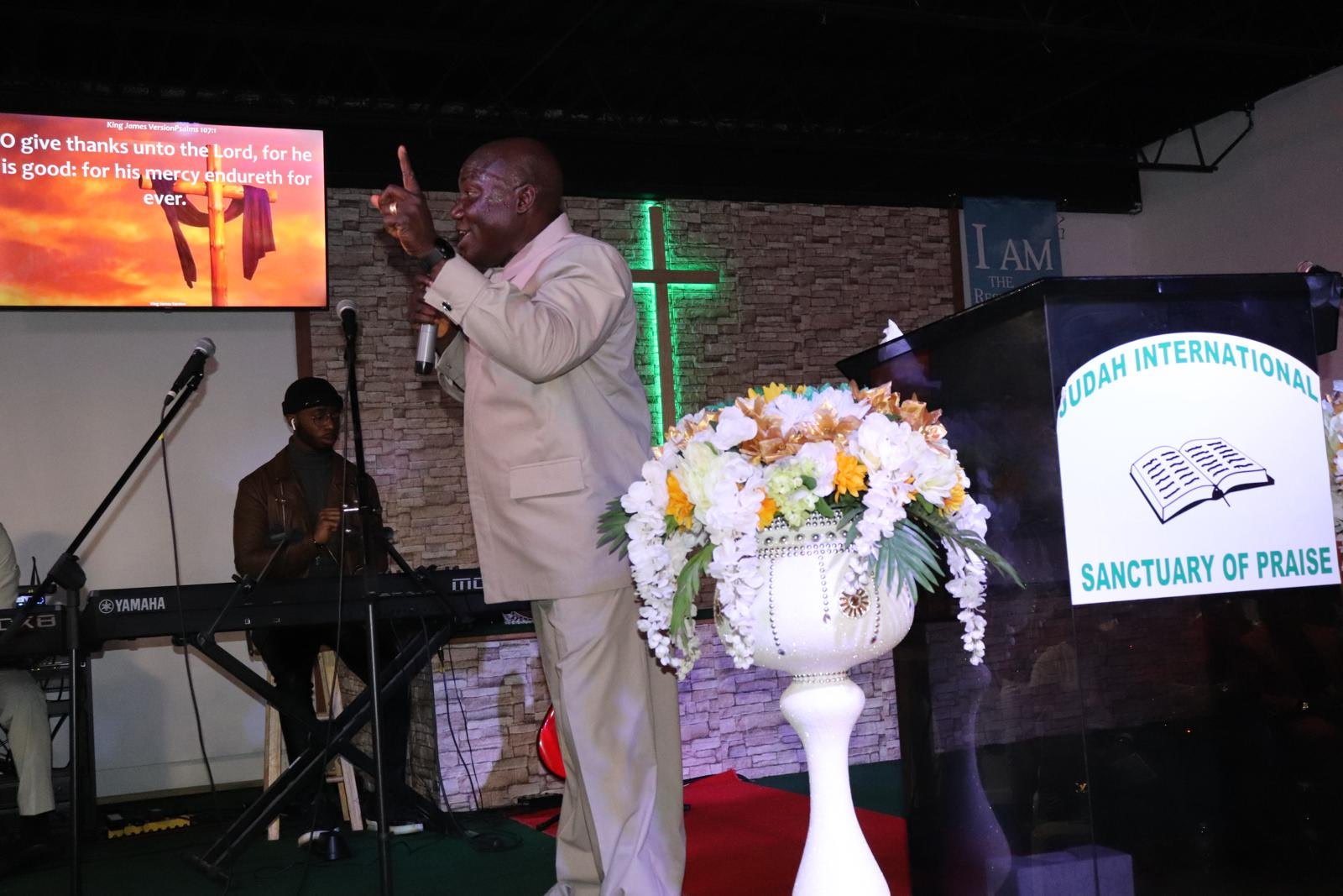 A man in a light-colored suit is speaking into a microphone while gesturing upward with his index finger at a church or religious gathering. Behind him, there is a woman playing a keyboard. The background features a brick wall with a lit green cross,