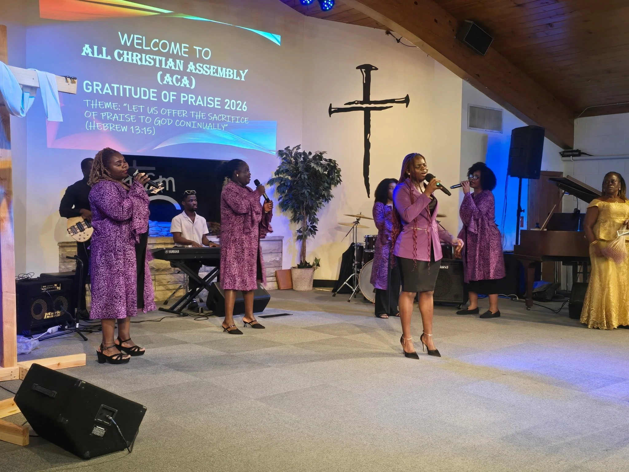 A group of people singing on stage inside a church, with a large cross on the wall behind them. The screen displays a welcome message for All Christian Assembly (ACA) and the theme for 2026 about offering praise to God.